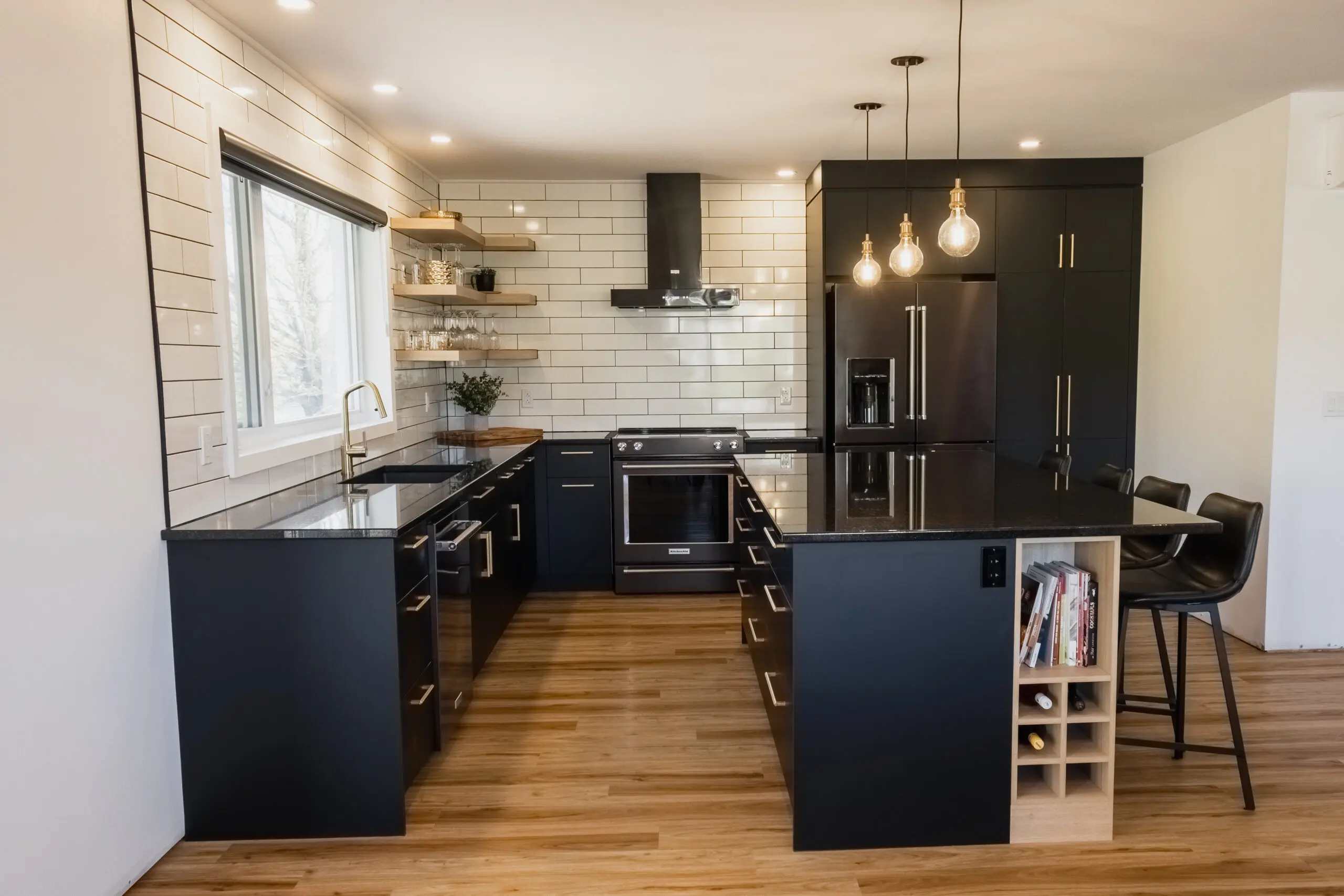 Cuisine moderne avec placards noirs, dosseret en carreaux de faïence blancs, appareils en acier inoxydable, sols en bois, étagères ouvertes, grande fenêtre, lampes suspendues au-dessus d'un îlot central avec tabourets de bar, et un casier à vin encastré.