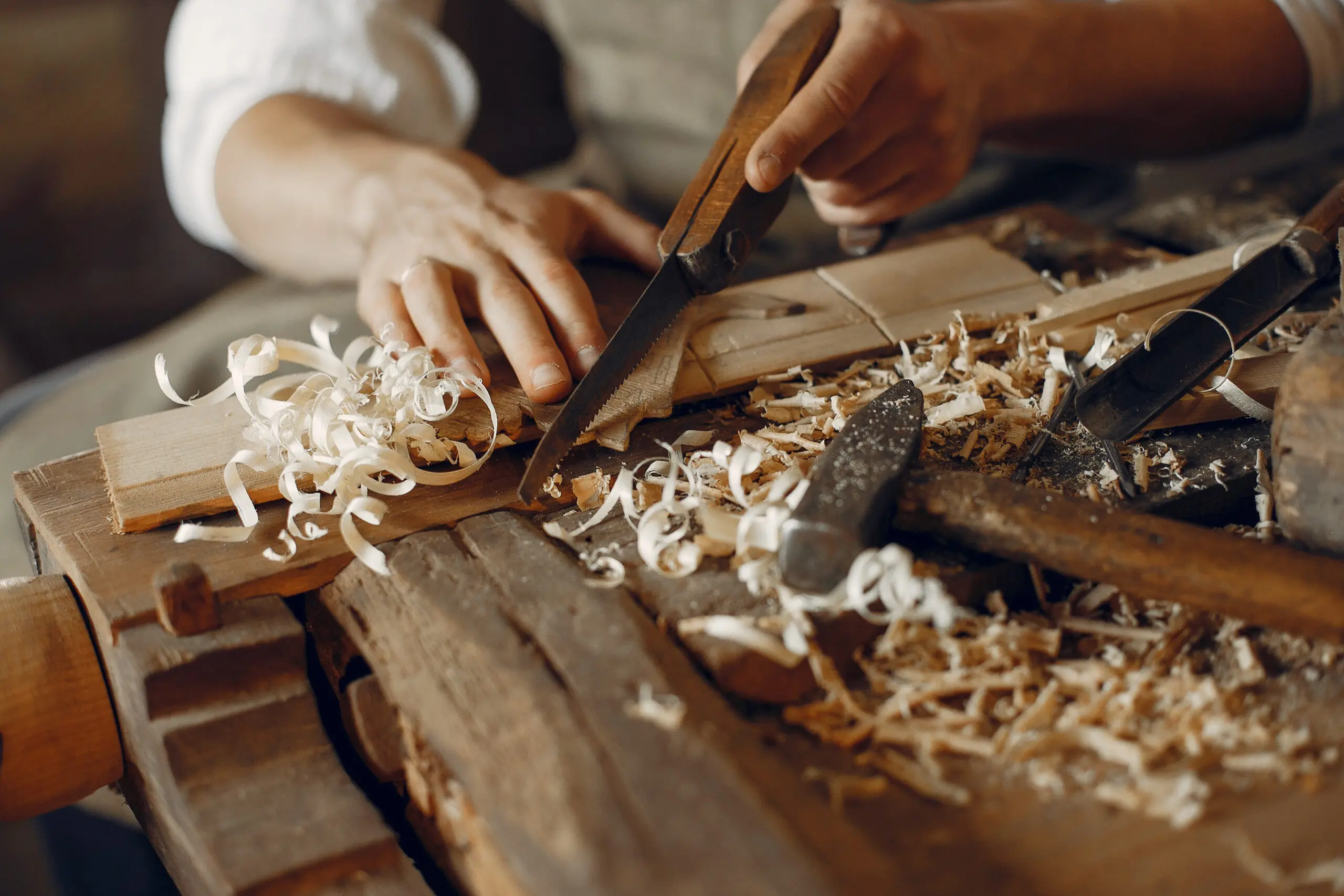 Une personne utilise un rabot manuel pour lisser une pièce de bois sur un établi, entouré de copeaux de bois et d'outils de travail du bois.
