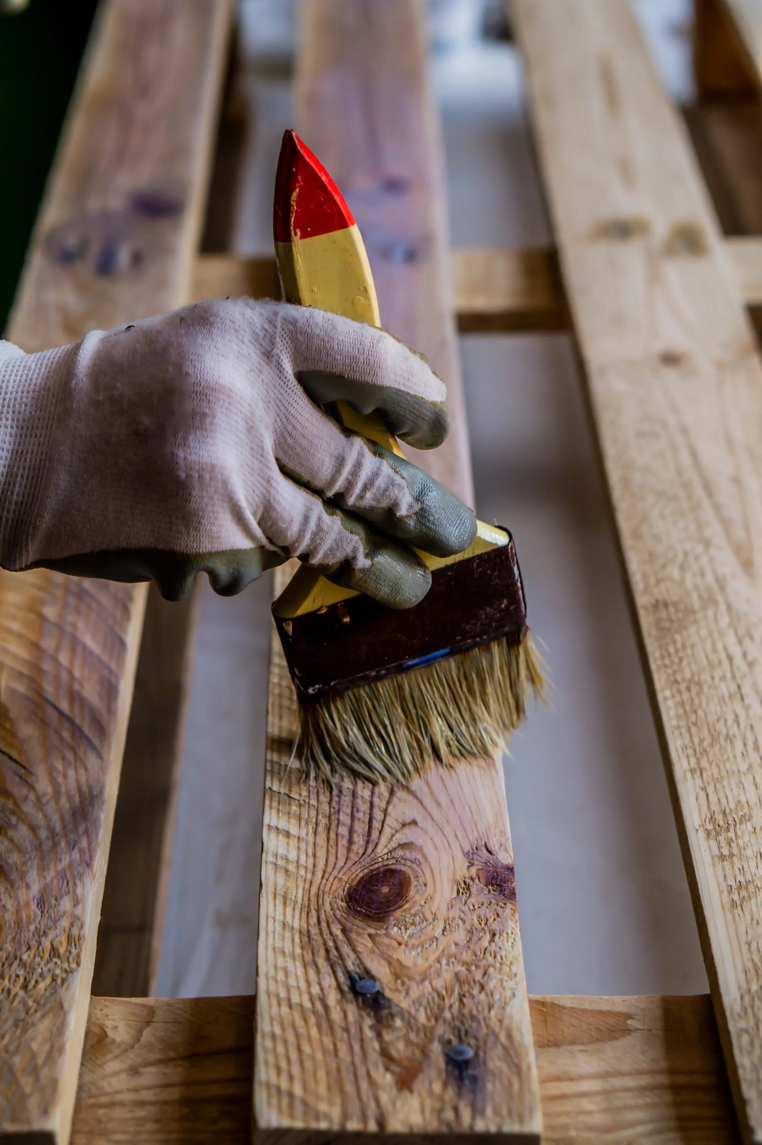 Une main gantée utilise un pinceau pour appliquer du vernis ou de la teinture sur une palette en bois, mettant en valeur le grain du bois et les nœuds.