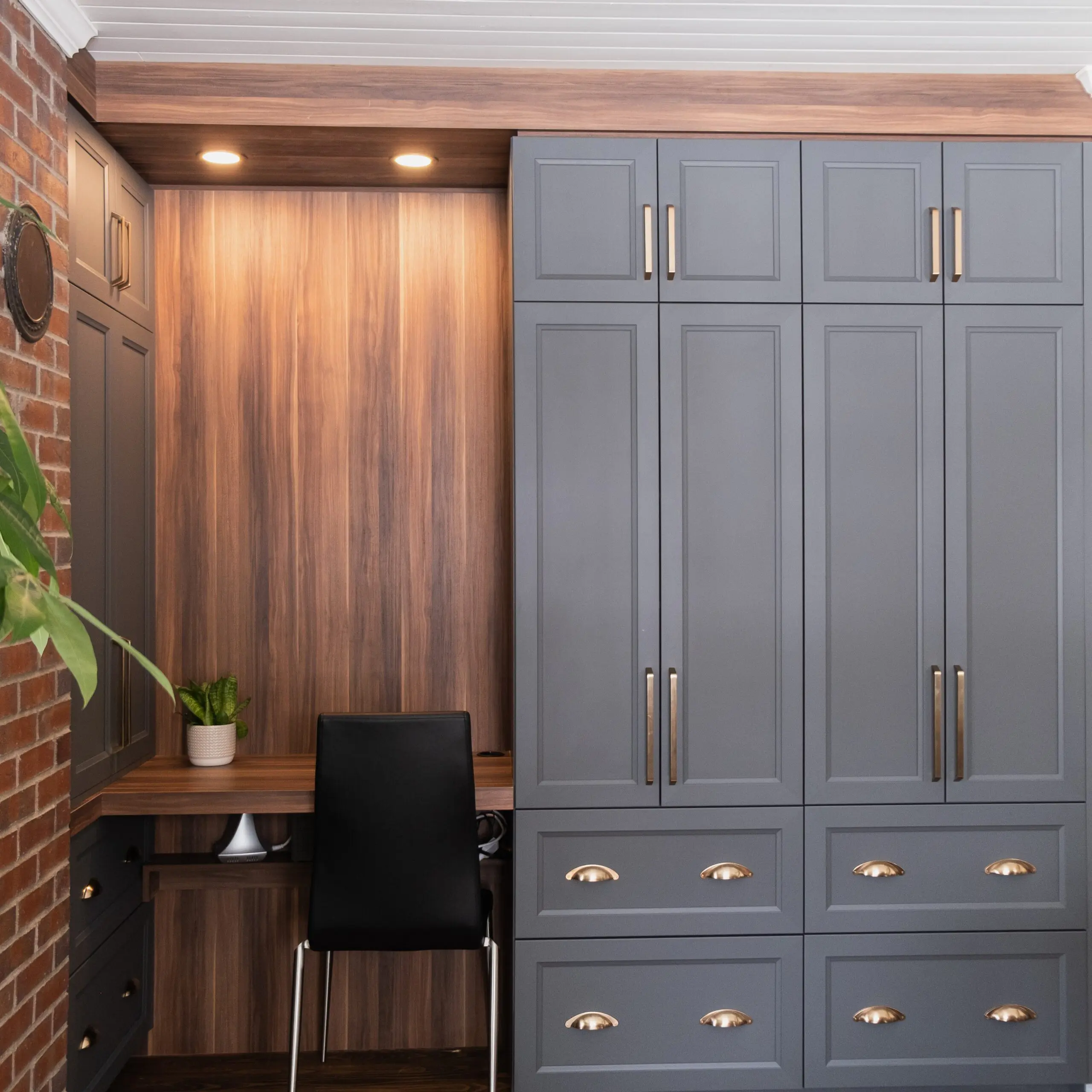 Bureau à domicile encastré avec un bureau en bois, des armoires grises, des poignées en laiton, une chaise noire, une plante en pot et des lumières encastrées au plafond, le tout contre un mur en briques et un plafond blanc.