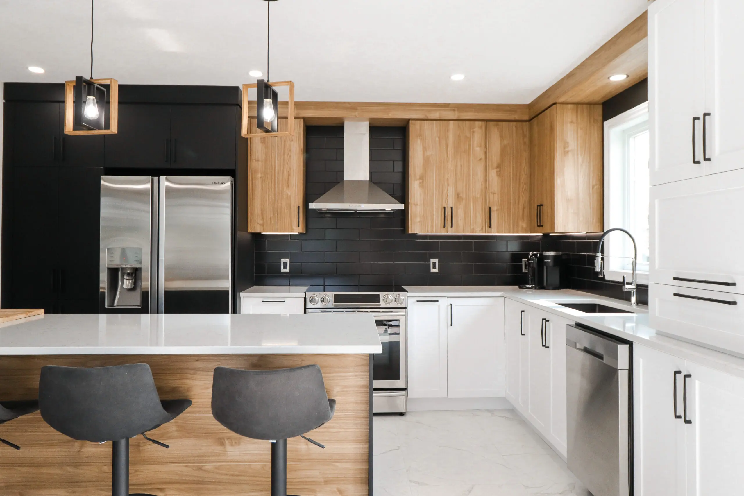 Cuisine moderne avec un mélange d'armoires en bois, noires et blanches, des appareils en acier inoxydable, un îlot central avec deux tabourets de bar sombres, des lampes suspendues et une fenêtre au-dessus de l'évier.