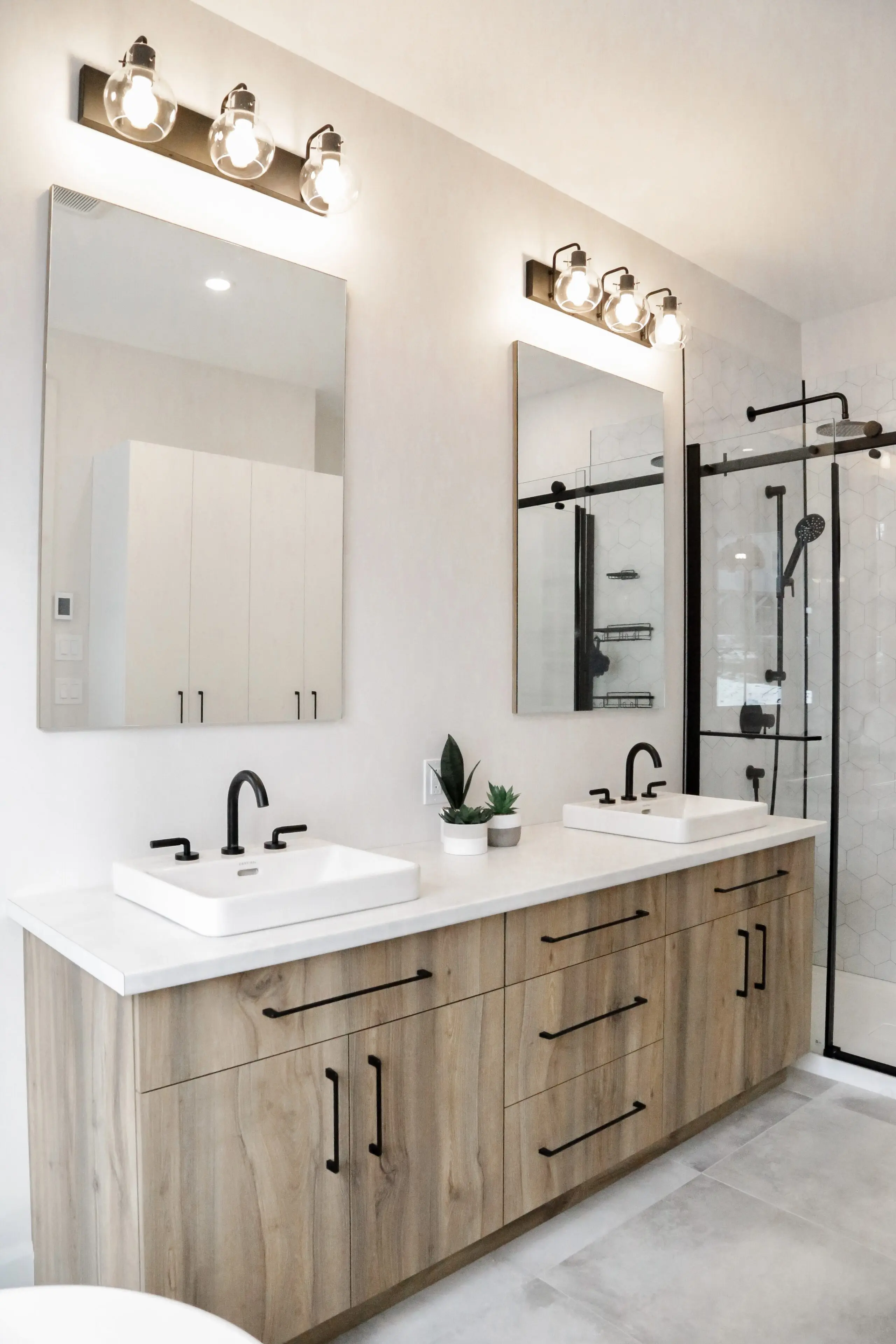 Salle de bains moderne avec un meuble double en bois, deux éviers carrés, des robinets noirs, deux grands miroirs avec des luminaires au-dessus, une petite plante en pot et une cabine de douche en verre avec des ferrures noires à l'arrière-plan.