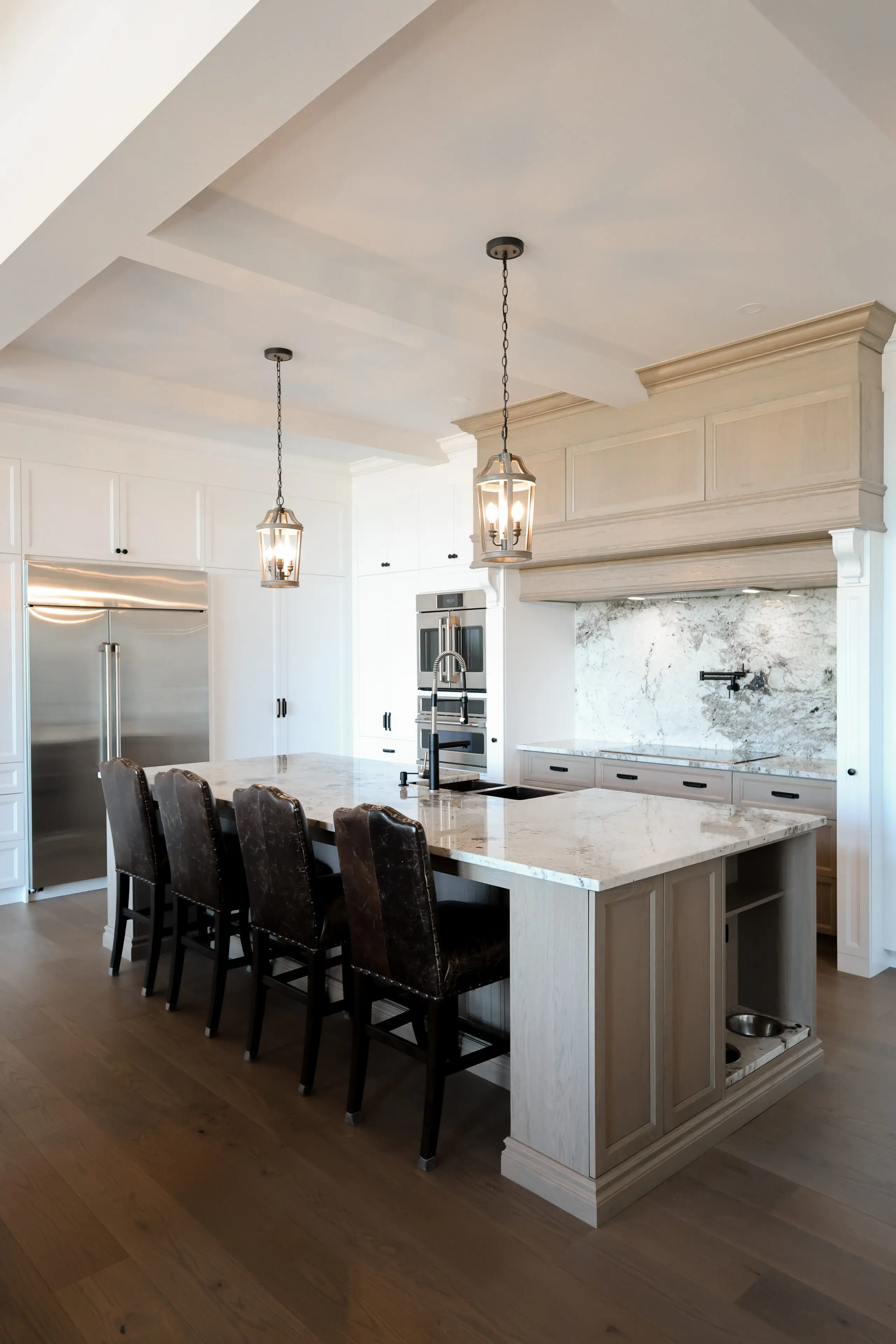Cuisine moderne avec un grand îlot doté d'un comptoir en marbre, quatre chaises sombres rembourrées, des lampes suspendues, des appareils en acier inoxydable intégrés et des planchers en bois clair.