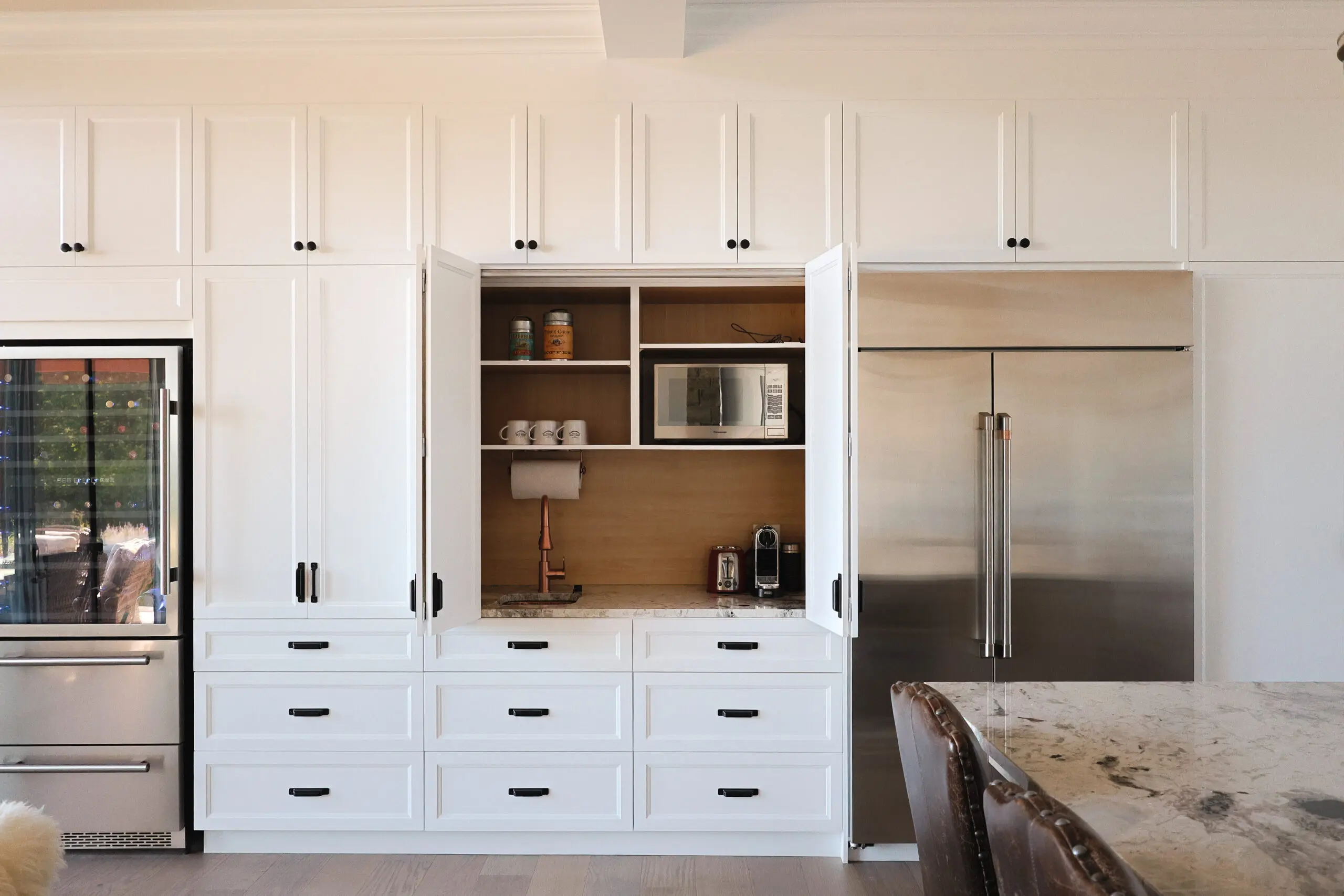 Cuisine moderne avec des armoires blanches, dont certaines s'ouvrent pour révéler des étagères avec un micro-ondes, des tasses et une cafetière. Un grand réfrigérateur en acier inoxydable se trouve à droite. Un comptoir en marbre et une partie d'une chaise sont visibles au premier plan.