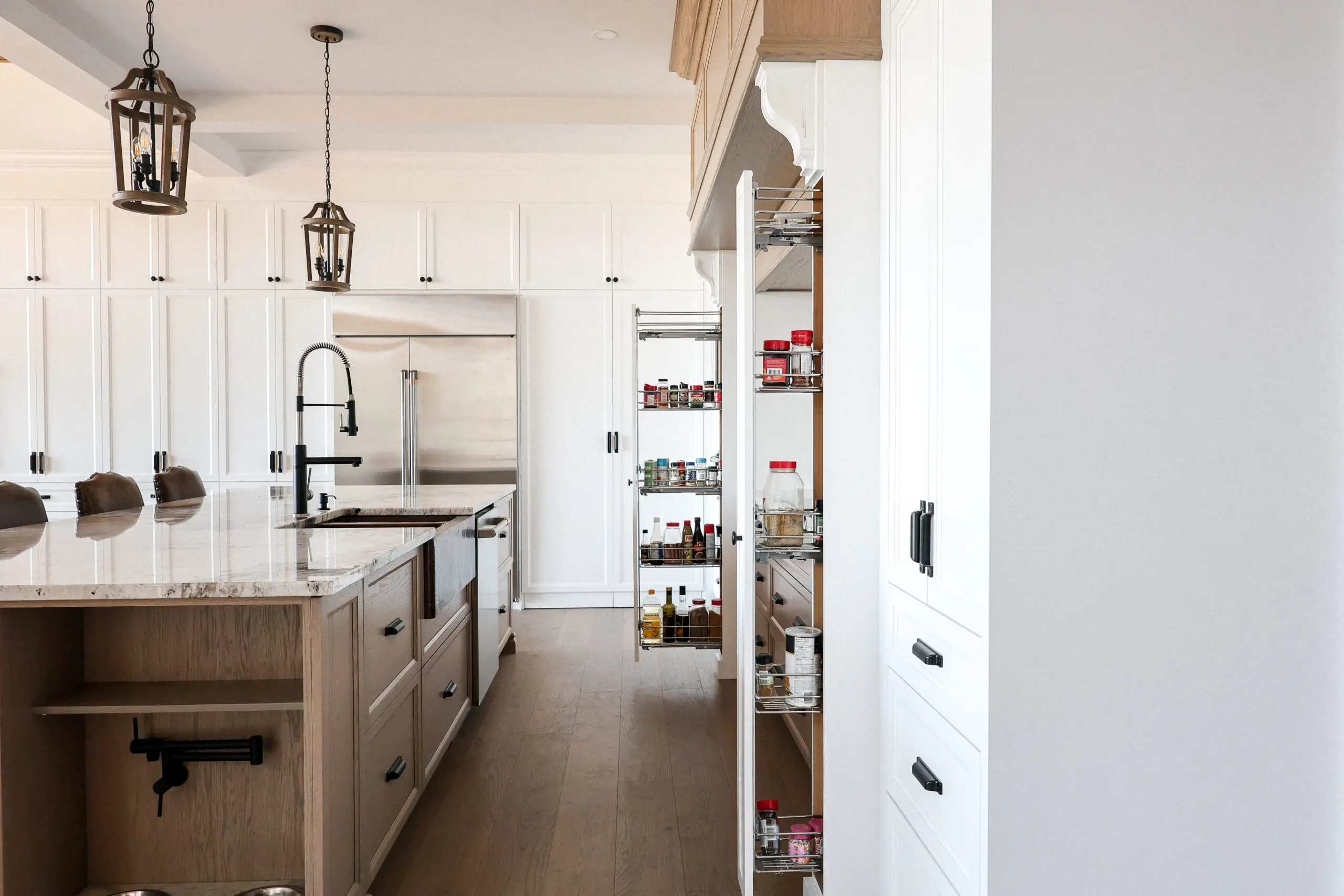 Une cuisine moderne comprend des armoires blanches, un grand îlot avec un évier intégré, des lampes suspendues et deux hautes étagères de garde-manger coulissantes remplies de divers produits alimentaires. La lumière naturelle éclaire cet espace spacieux et bien organisé.