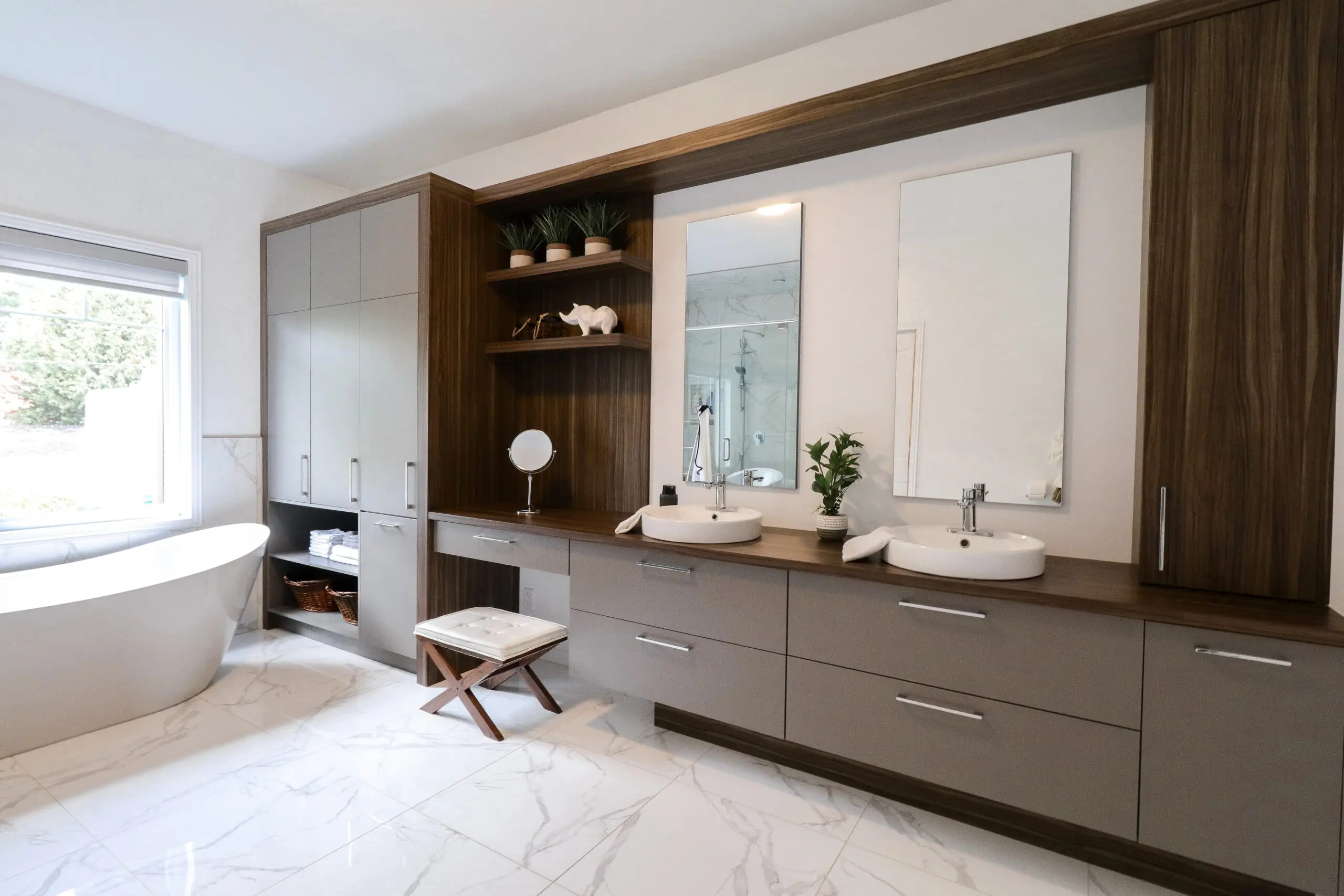 Salle de bains moderne avec de grandes armoires en bois, deux lavabos avec miroirs, une baignoire autoportante près d'une fenêtre, un sol en carreaux de marbre, et des plantes et accessoires décoratifs sur des étagères ouvertes.