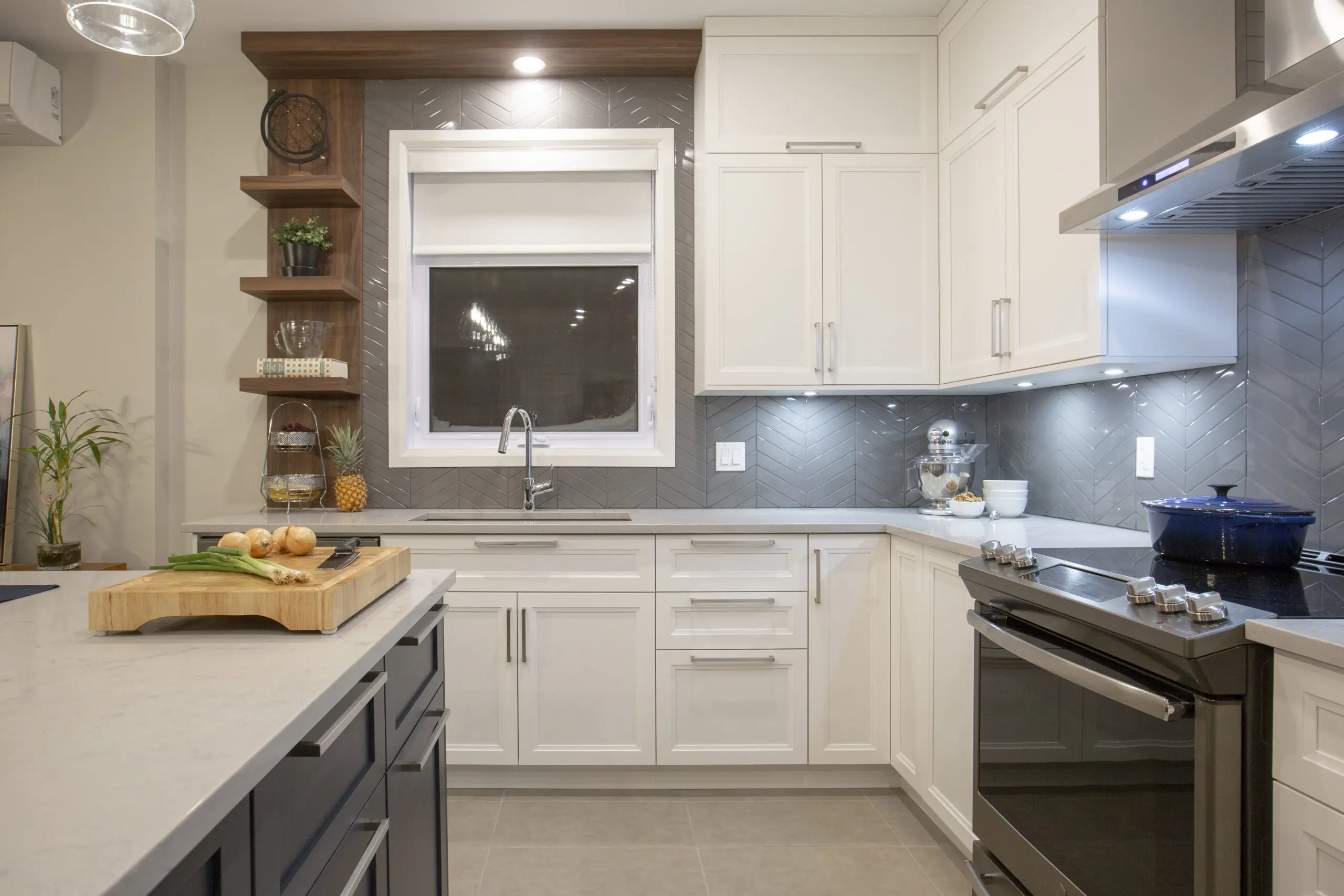 Cuisine moderne avec des armoires blanches, un dosseret gris, des appareils électroménagers en acier inoxydable et des étagères ouvertes en bois. Un îlot avec une planche à découper et du pain se trouve au premier plan, et un éclairage sous l'armoire illumine les plans de travail.