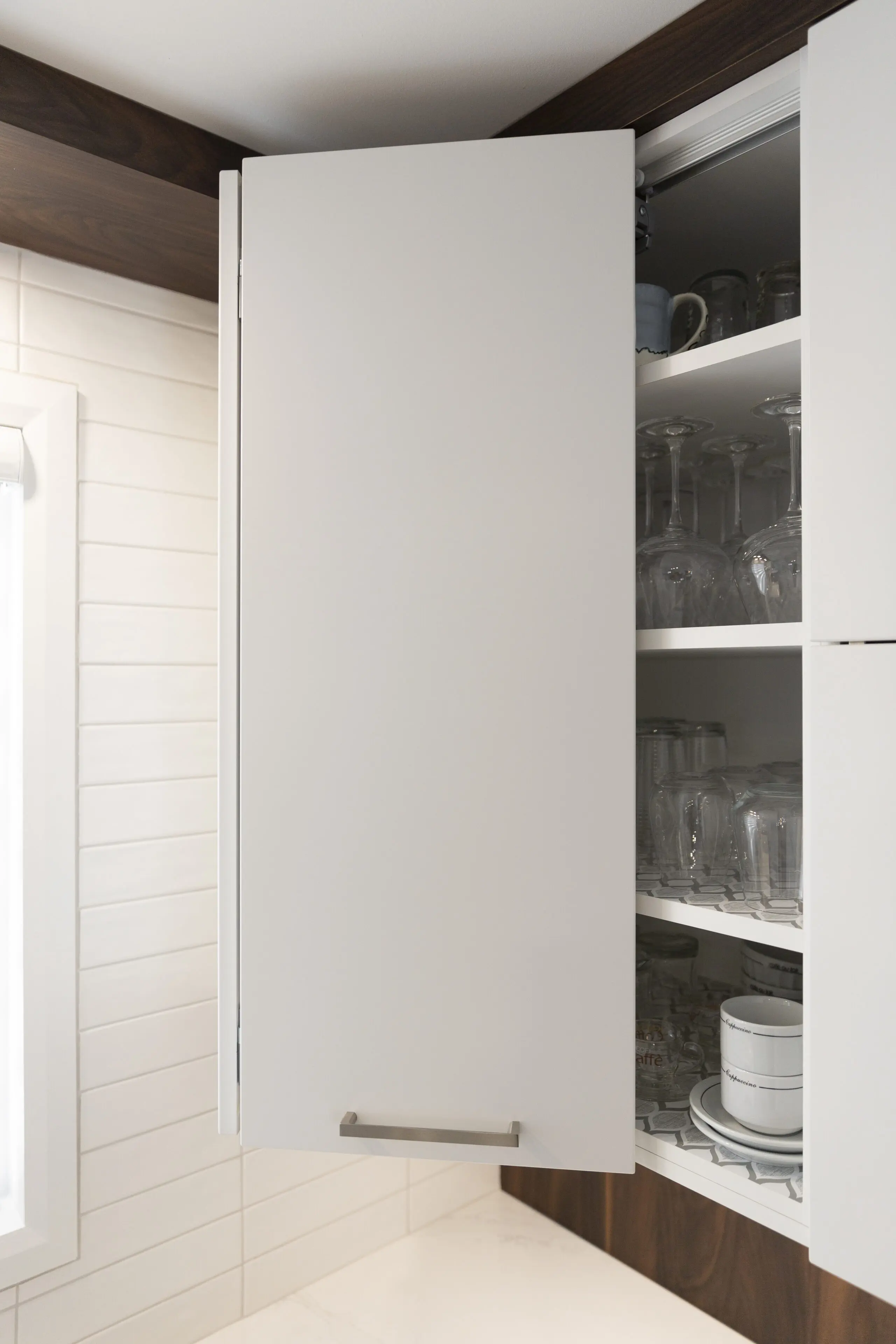 Un meuble de cuisine blanc dont la porte est partiellement ouverte, révélant des étagères contenant des verres, des tasses et des bols. Le meuble est monté sur un mur carrelé à côté d'une fenêtre.