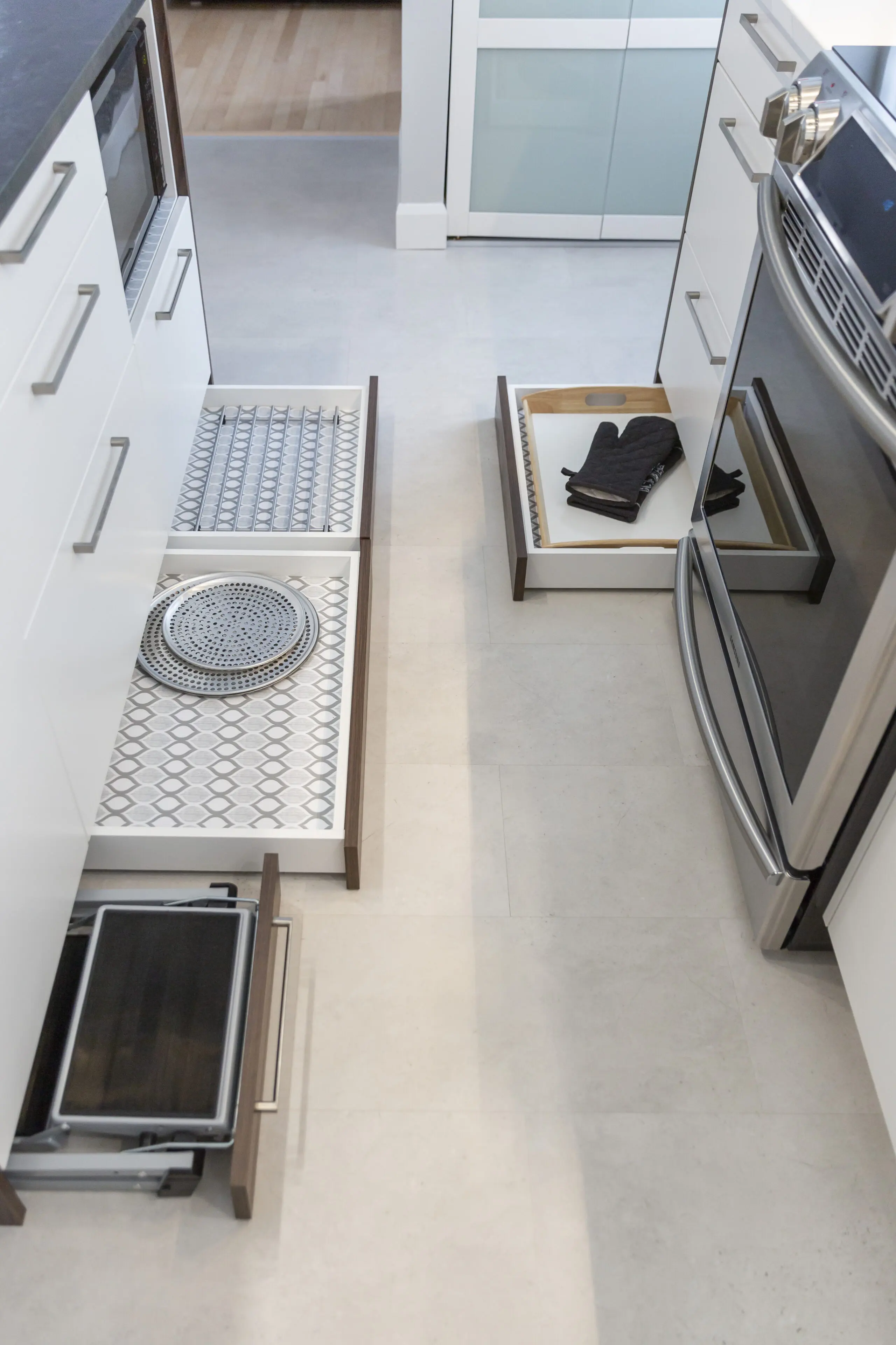 Des tiroirs de cuisine ouverts révèlent un rangement organisé pour les plateaux, les gants de cuisine et une grille de séchage à côté d'un four en acier inoxydable. Les tiroirs sont bien rangés dans une cuisine moderne et blanche.