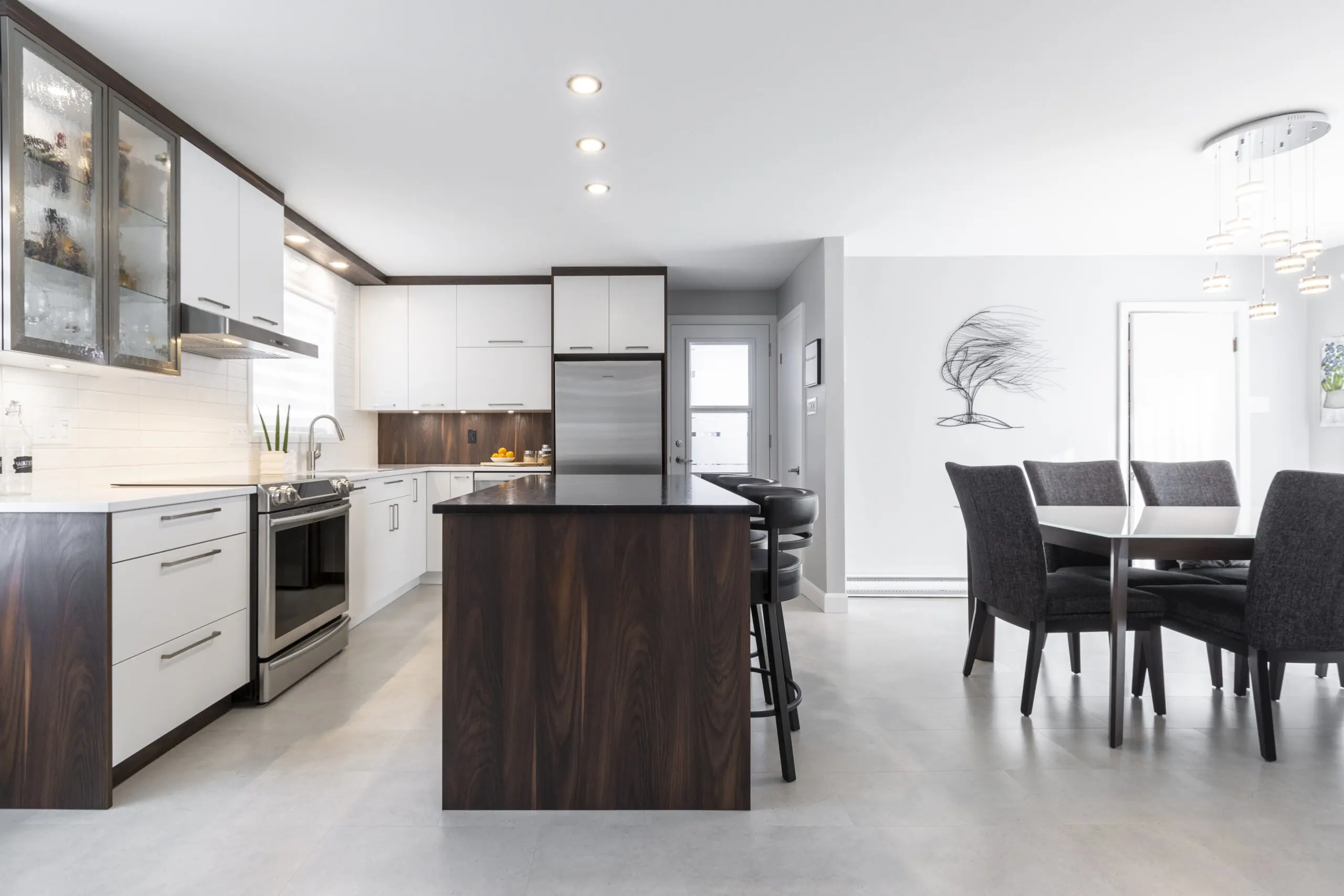 Cuisine et salle à manger modernes avec des armoires blanches, un îlot en bois foncé, des appareils électroménagers en acier inoxydable et une table à manger de six places. Des suspensions sont suspendues au-dessus de la table et une œuvre d'art murale représentant un arbre décore la pièce lumineuse.