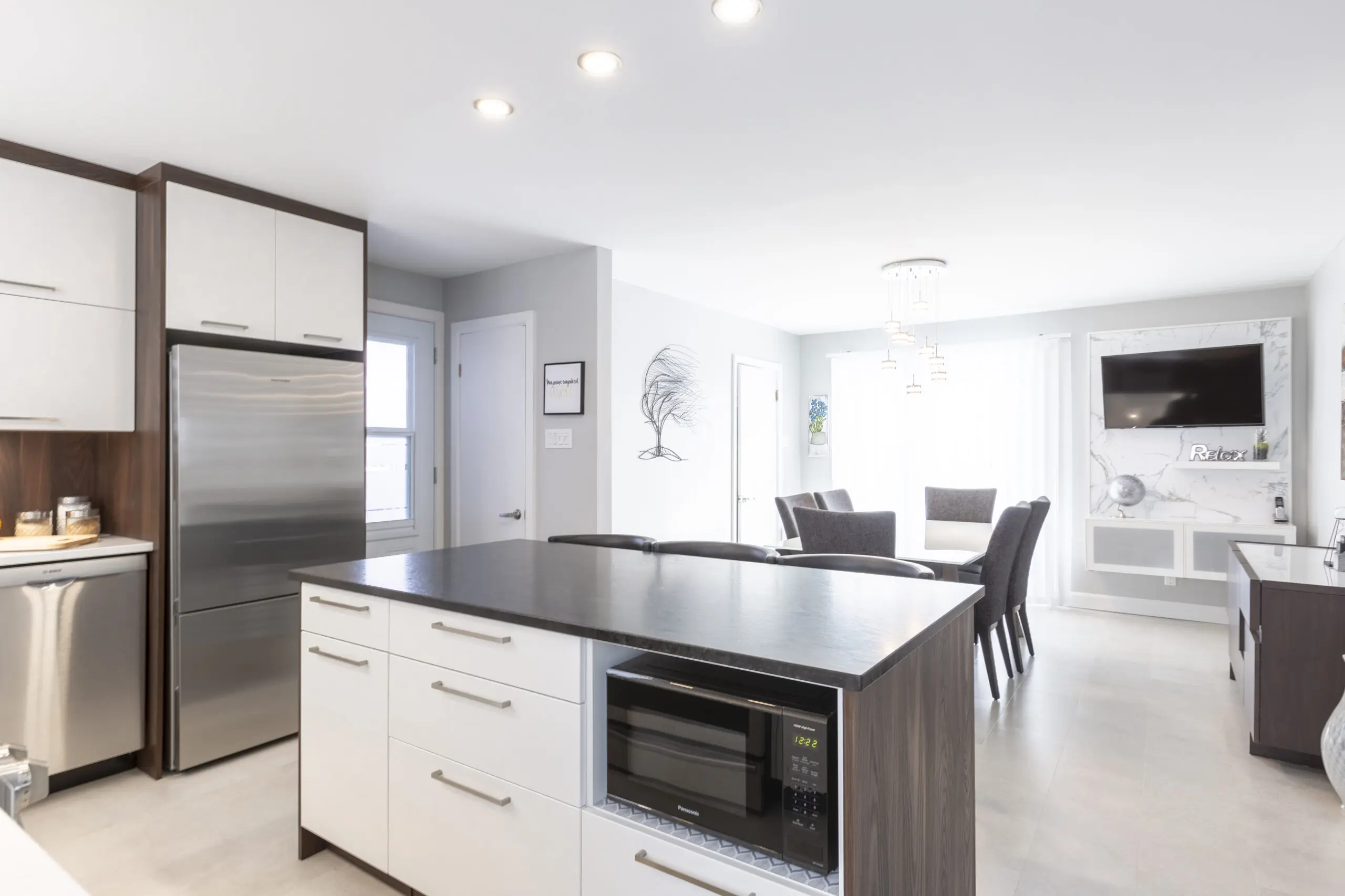 Cuisine et salle à manger modernes avec des armoires blanches, des appareils en acier inoxydable, un grand îlot avec un micro-ondes intégré, et une table à manger avec six chaises près de grandes fenêtres laissant entrer la lumière naturelle.