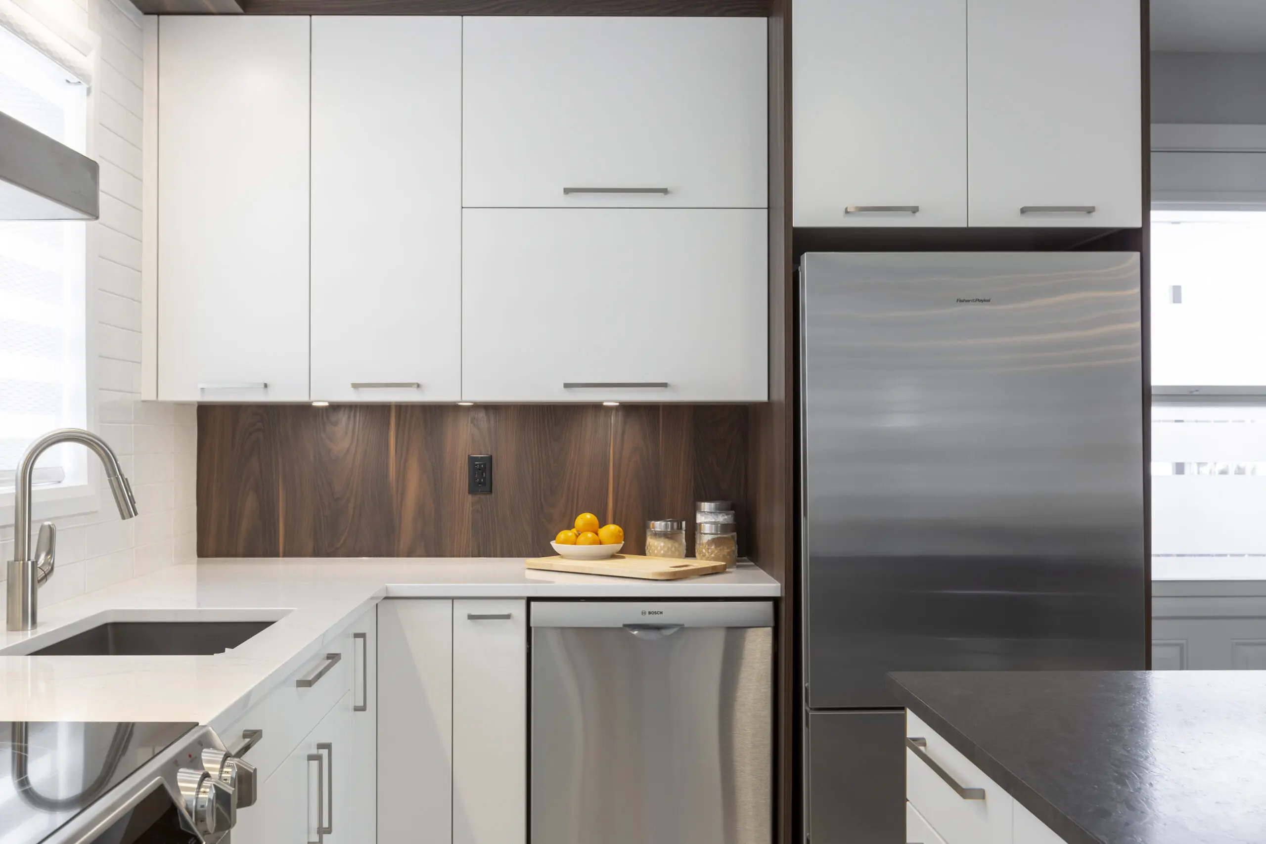 Cuisine moderne avec des armoires blanches, un réfrigérateur et un lave-vaisselle en acier inoxydable, un évier et un dosseret en bois. Un plateau rond en bois contenant des oranges et des bocaux est posé sur le comptoir près du réfrigérateur.