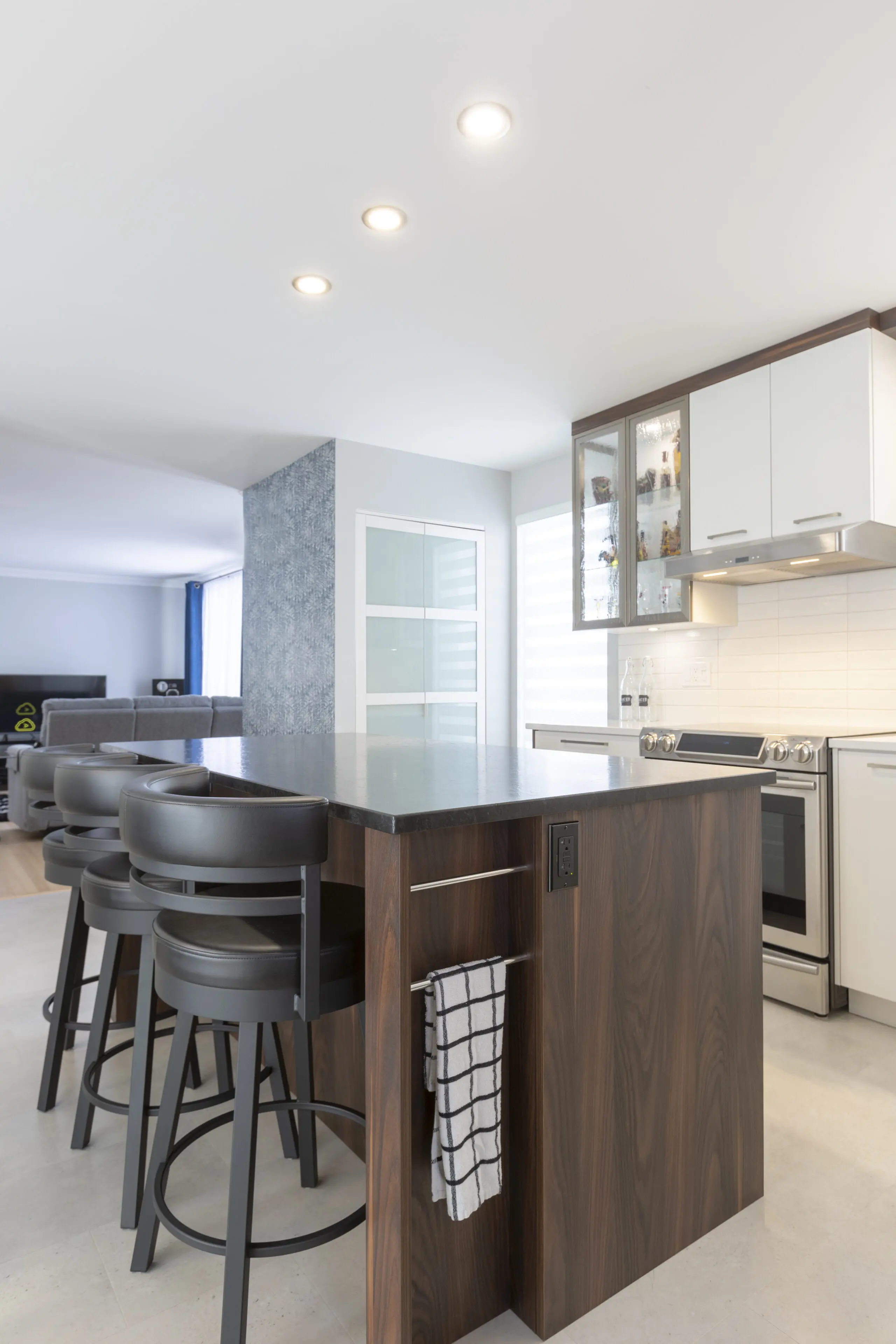 Cuisine moderne avec un îlot en bois foncé, quatre tabourets noirs, une serviette rayée suspendue sur le côté, des placards blancs, des appareils électroménagers en acier inoxydable et un éclairage zénithal lumineux.