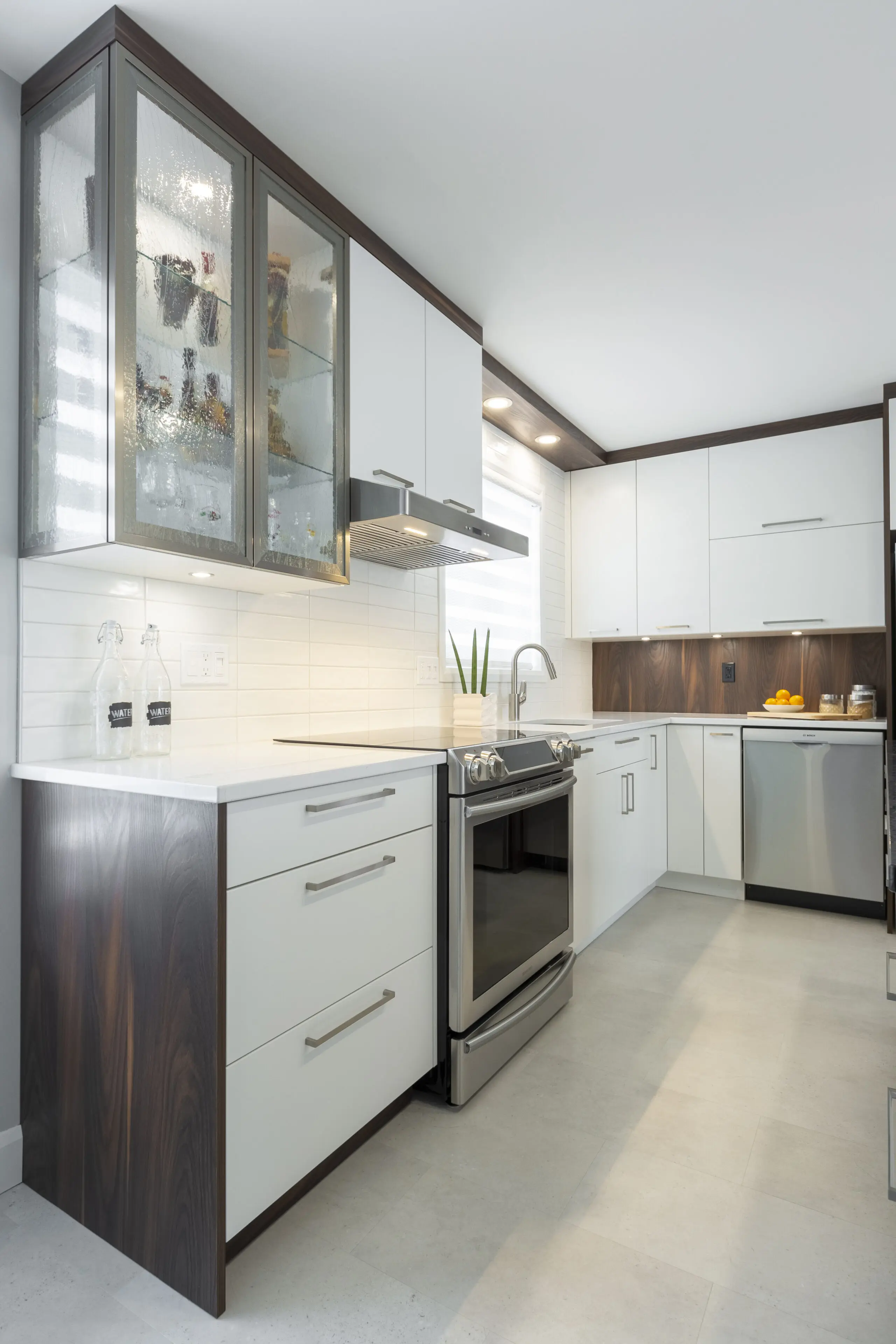 Une cuisine moderne avec des armoires blanches, des appareils en acier inoxydable, des armoires supérieures en verre, un four encastré, un décor minimaliste, une petite plante en pot et un éclairage sous l'armoire.