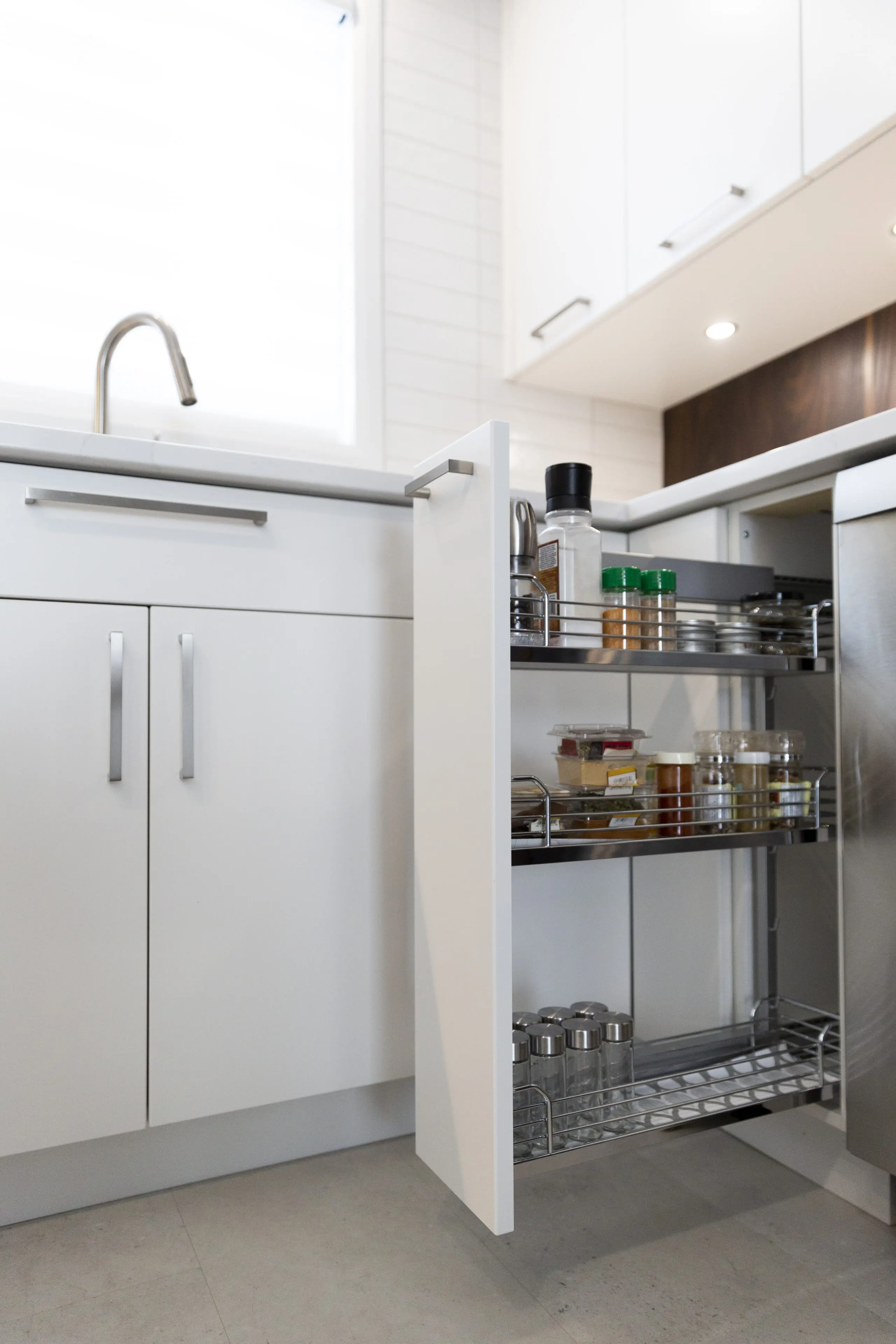 Une cuisine blanche moderne avec une armoire à tiroirs ouverte, présentant des épices et des condiments bien rangés sur des étagères en métal. Le plan de travail, l'évier et les armoires sont propres et minimalistes.
