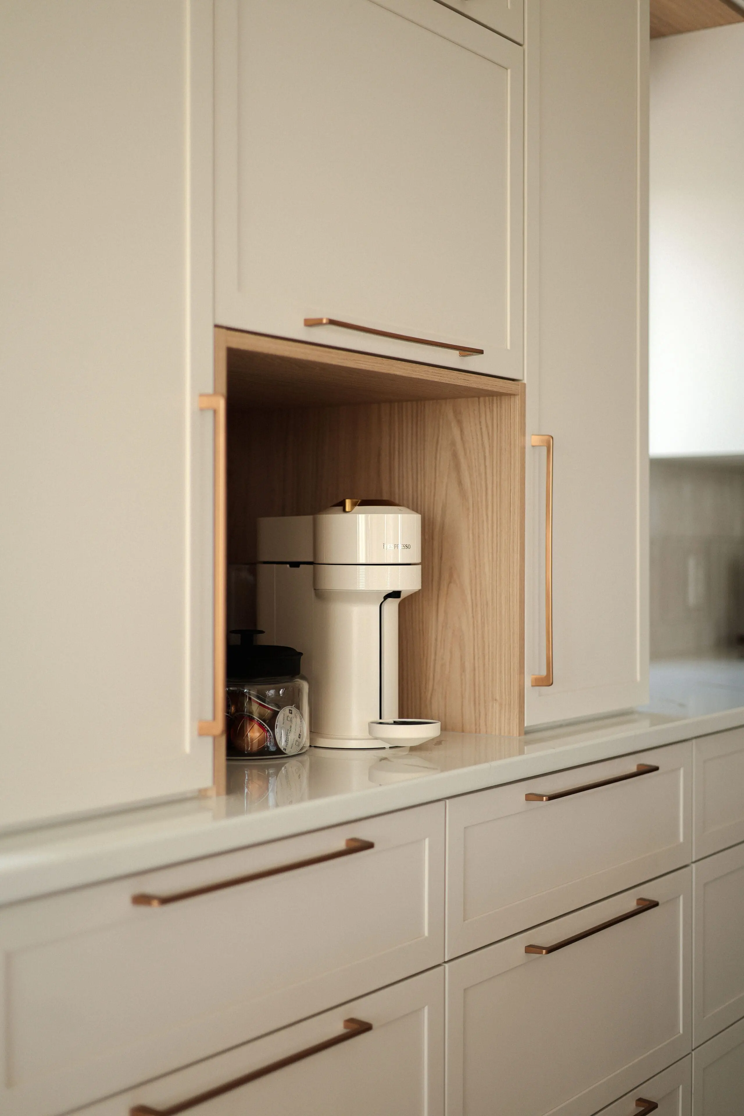 Un coin cuisine moderne avec une cafetière encastrée, un pot en verre contenant des dosettes de café et des armoires de couleur claire avec des poignées en cuivre élégantes.
