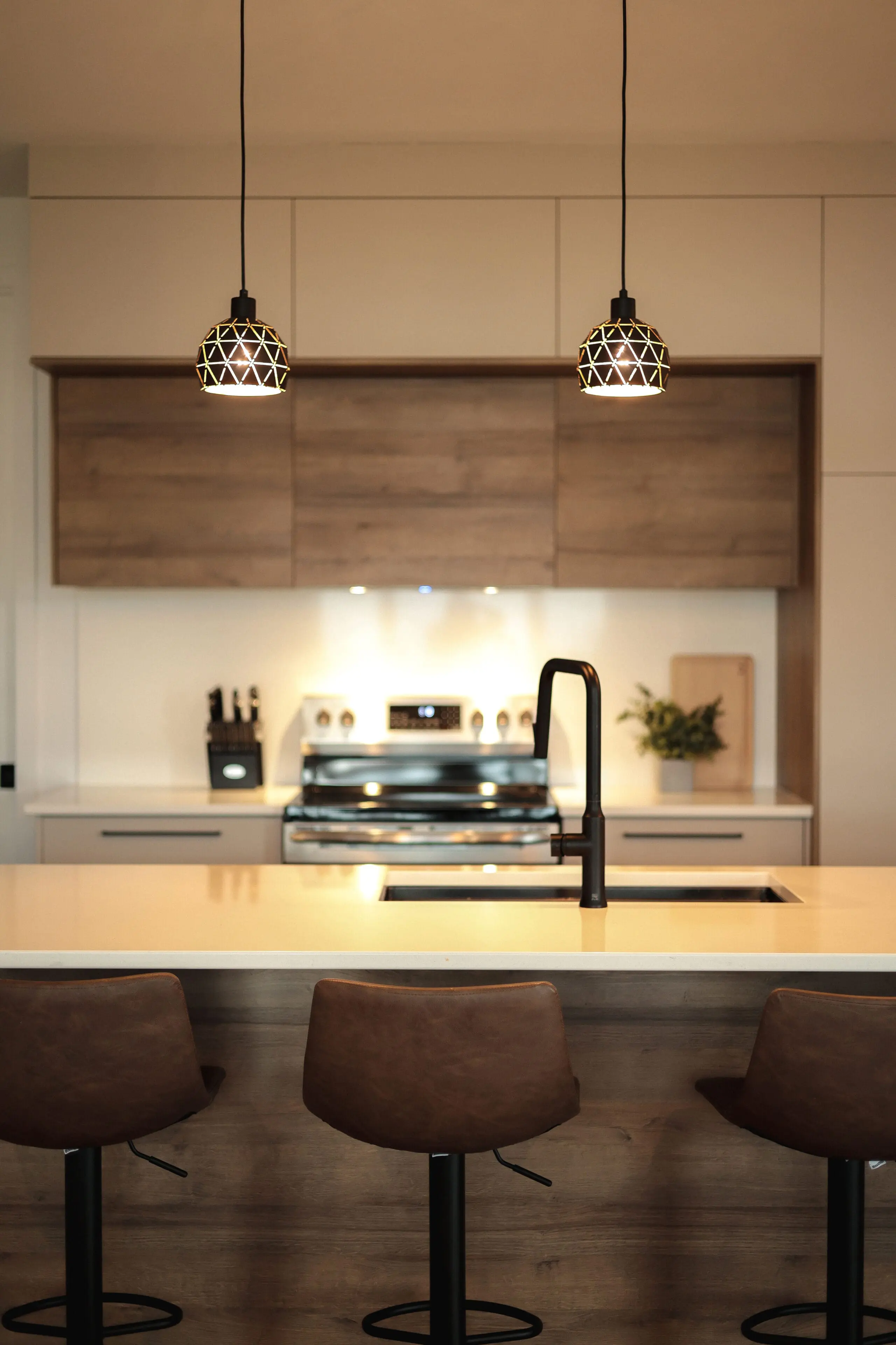 Cuisine moderne avec des armoires en bois, un îlot central avec trois tabourets de bar marron, un robinet noir, des appareils en acier inoxydable et deux lampes suspendues géométriques suspendues au-dessus du comptoir.