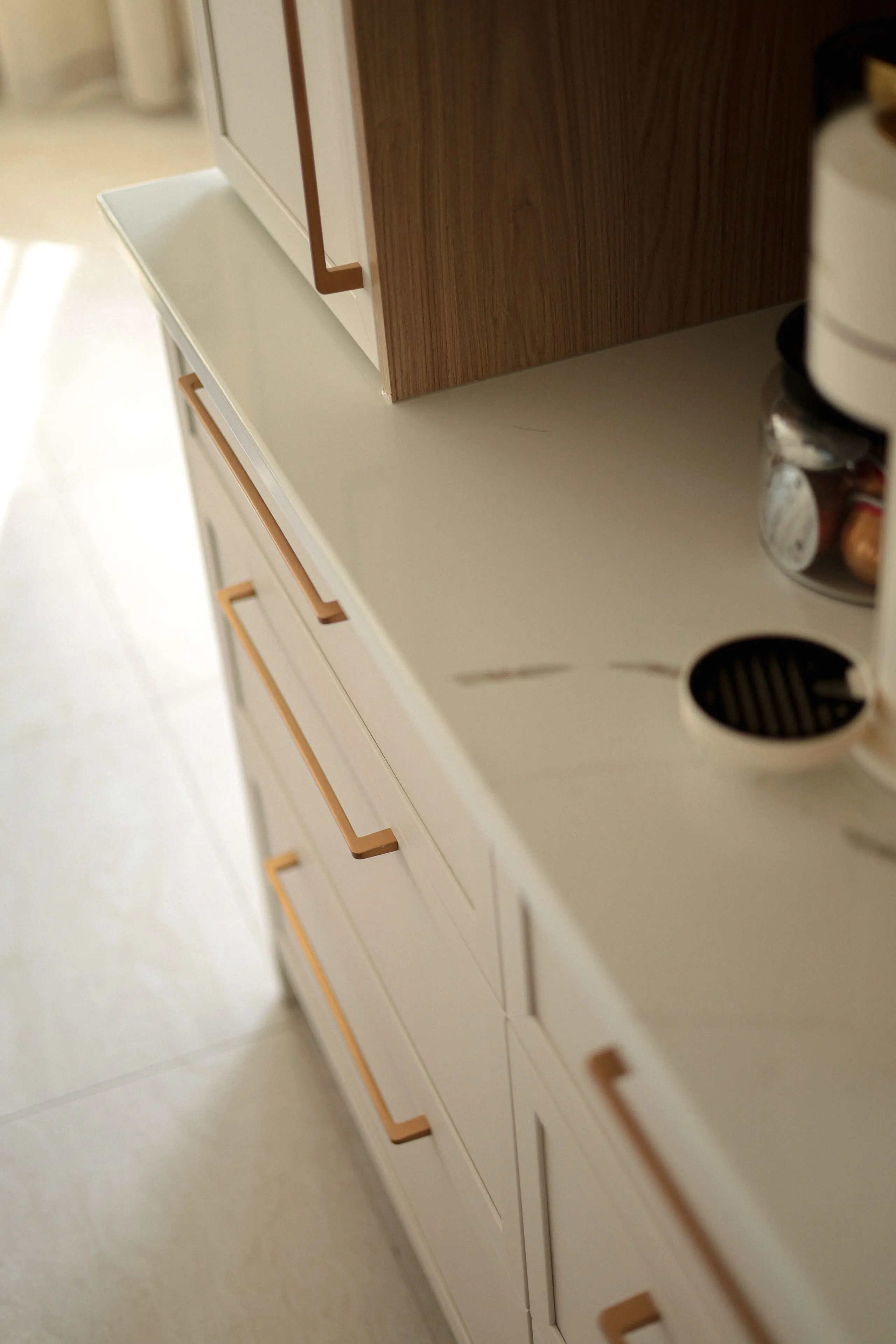 Gros plan sur un comptoir de cuisine moderne avec des meubles blancs et des poignées dorées, avec une surface en marbre et une machine à café avec un pot de capsules de café.