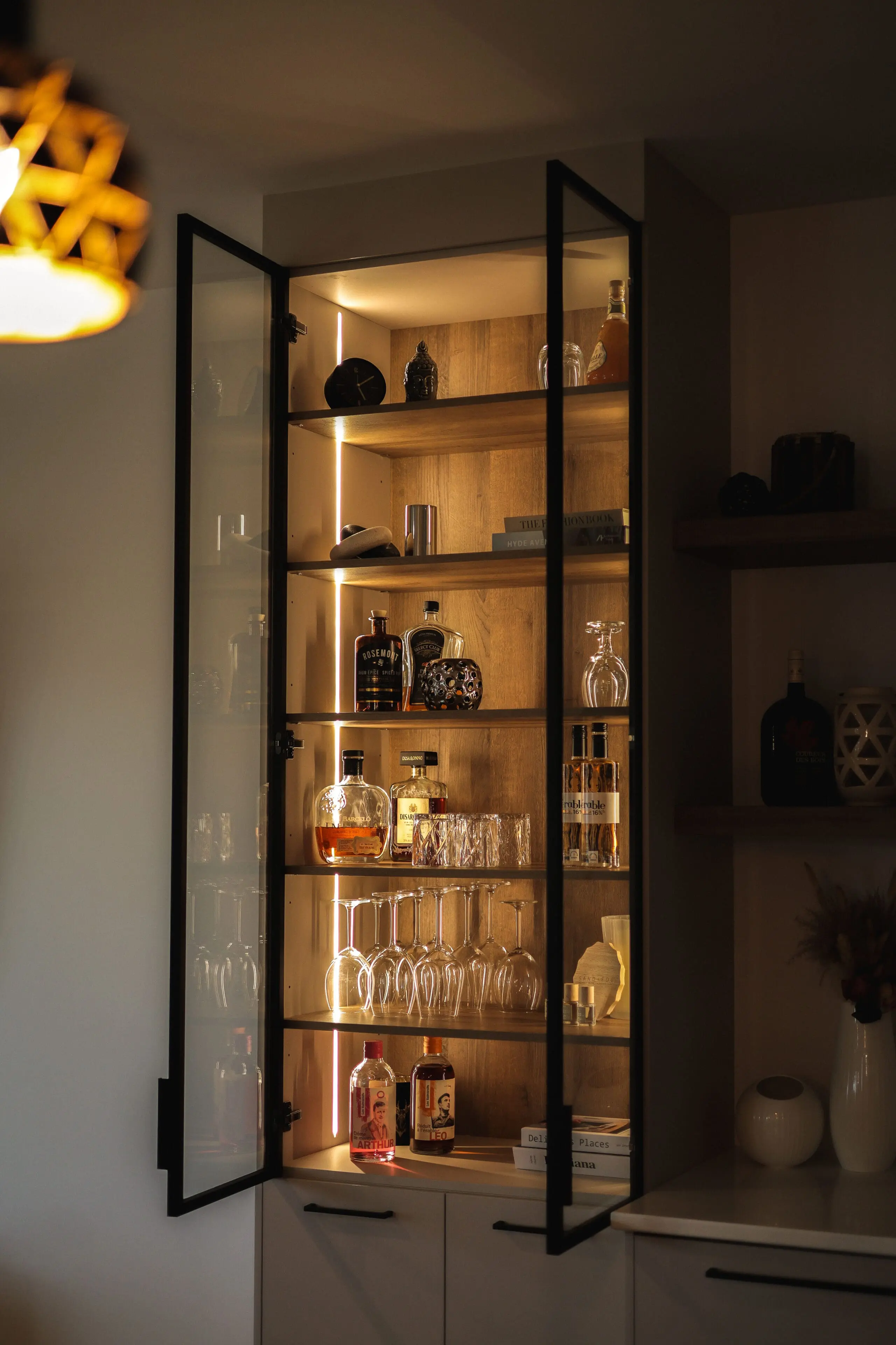 Une armoire vitrée avec des étagères présentant diverses bouteilles d'alcool, de la verrerie et des carafes, le tout éclairé par une lumière intérieure chaude. Certaines bouteilles et certains verres sont bien rangés sur les étagères.