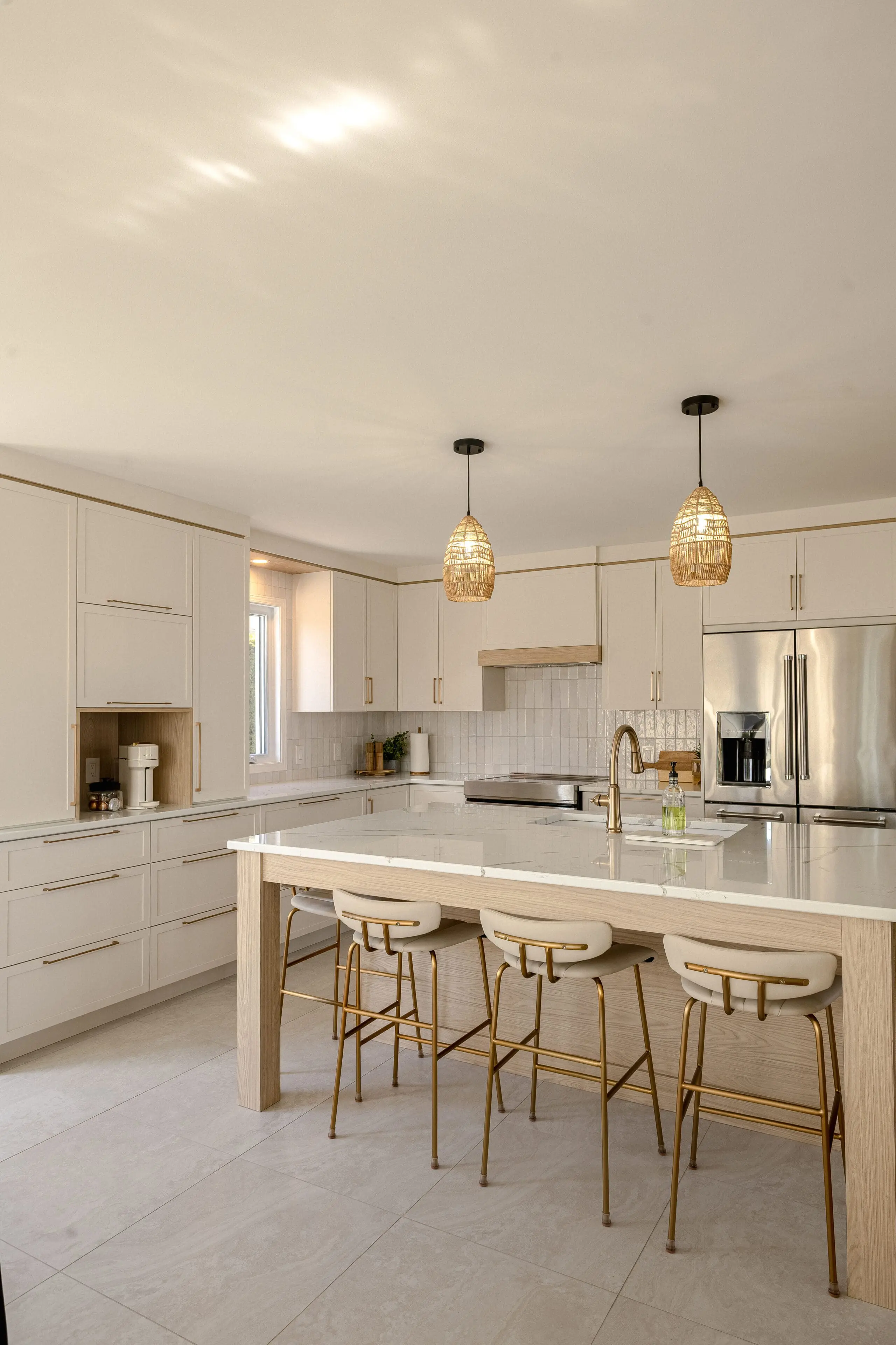 Cuisine moderne avec des armoires en bois clair, un grand îlot avec un comptoir blanc, trois tabourets de bar rembourrés, des appareils électroménagers en acier inoxydable et deux lampes suspendues tressées suspendues au-dessus de l'îlot. La lumière naturelle remplit l'espace.