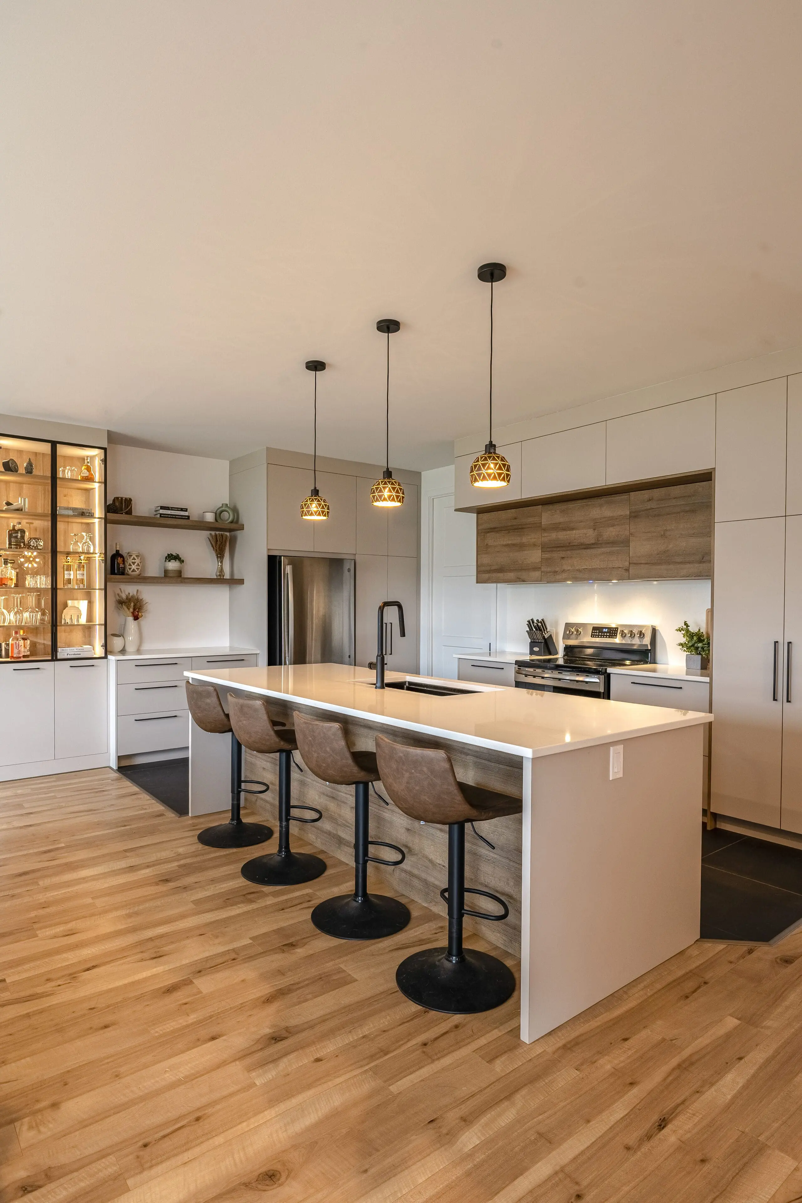 Cuisine moderne avec des planchers en bois clair, un grand îlot blanc avec quatre tabourets de bar bruns, des lampes suspendues au-dessus, des appareils encastrés, des armoires légères et des étagères ouvertes présentant de la vaisselle et de la décoration.