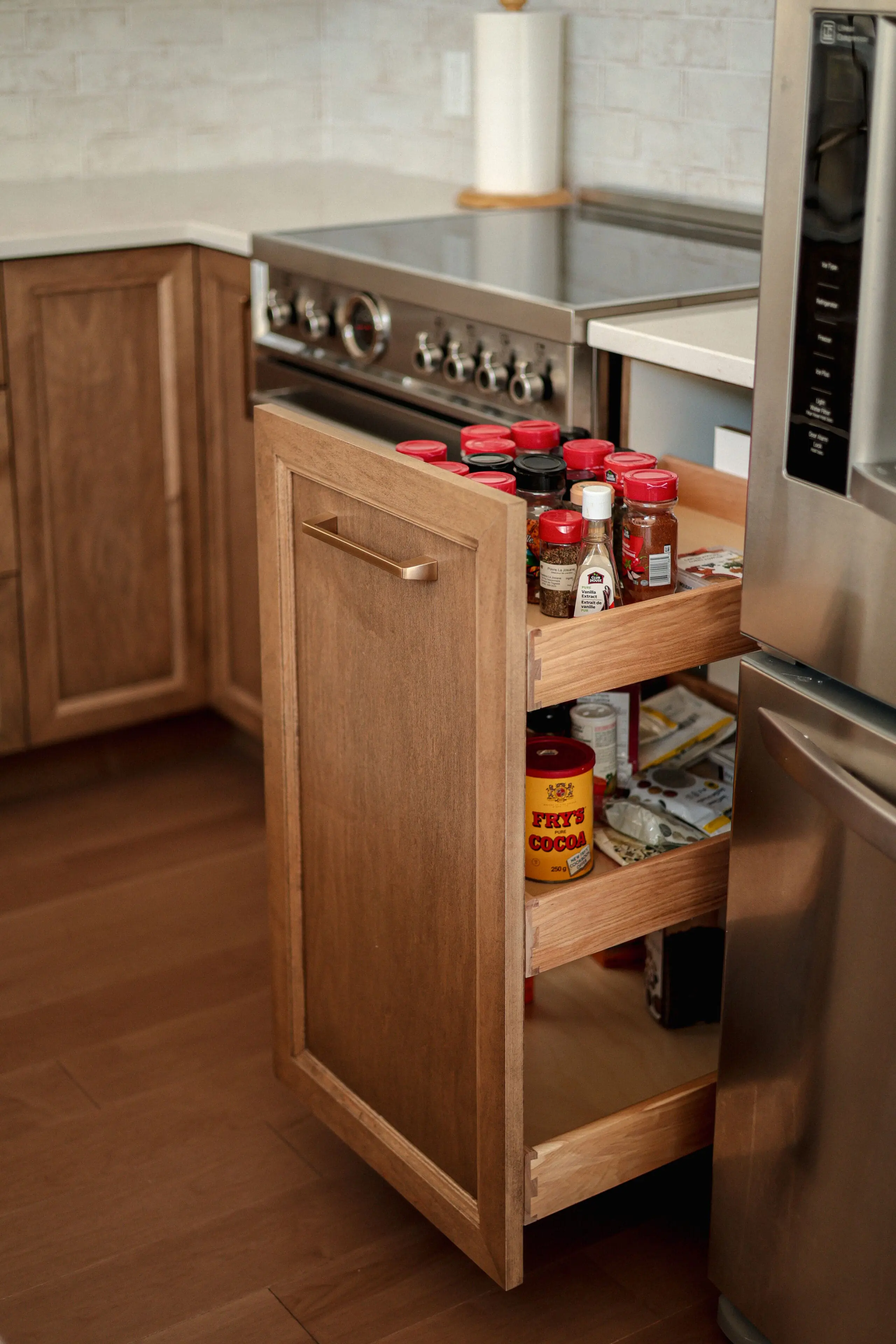 Dans une cuisine, un tiroir de garde-manger en bois est ouvert, révélant des étagères remplies de pots d'épices, de sauces et de conserves à côté d'une cuisinière et d'un réfrigérateur.