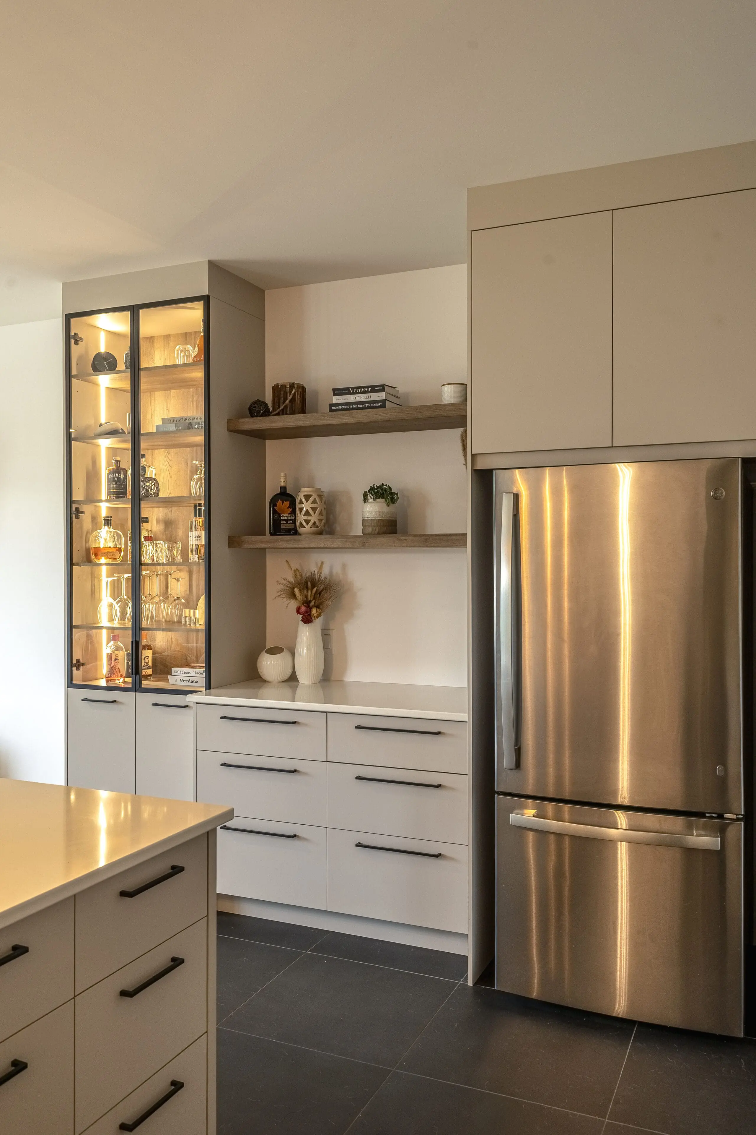 Cuisine moderne avec des armoires claires, un réfrigérateur en acier inoxydable, un meuble bar à façade vitrée avec des étagères éclairées contenant des bouteilles et des verres, des étagères ouvertes en bois avec des objets décoratifs, et un sol en carrelage foncé.