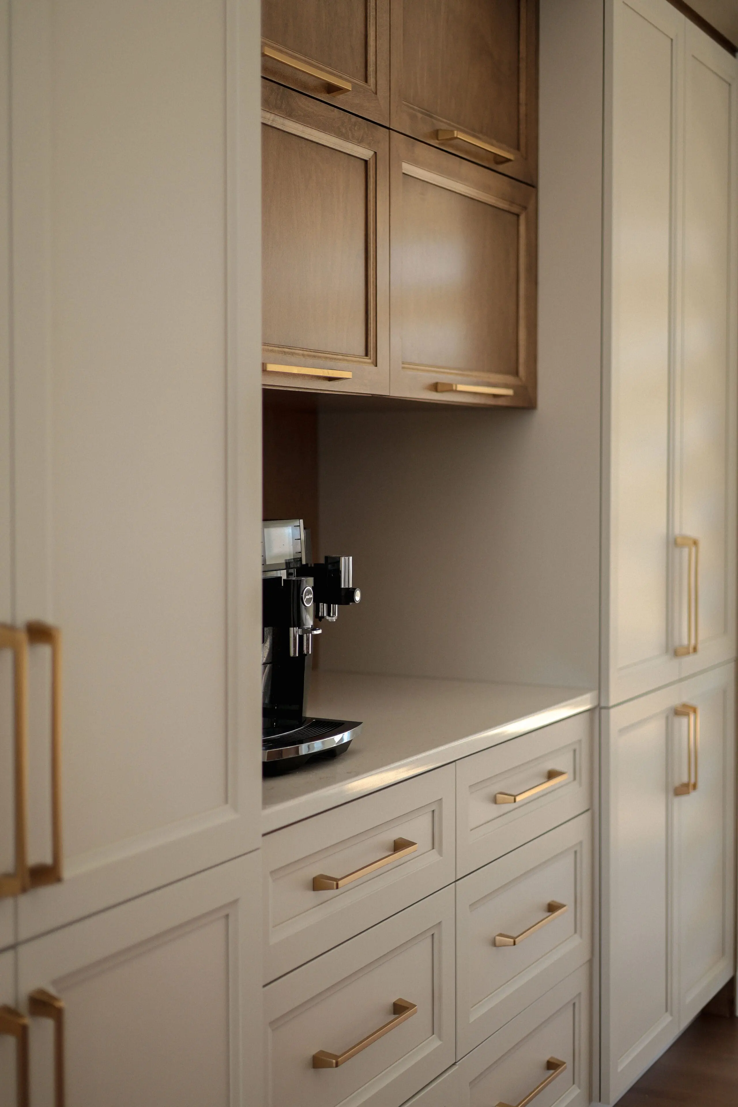 Machine à café encastrée dans un coin cuisine moderne avec des armoires beige clair, des poignées en laiton et des armoires supérieures en bois. L'espace présente des lignes épurées et un design minimaliste.