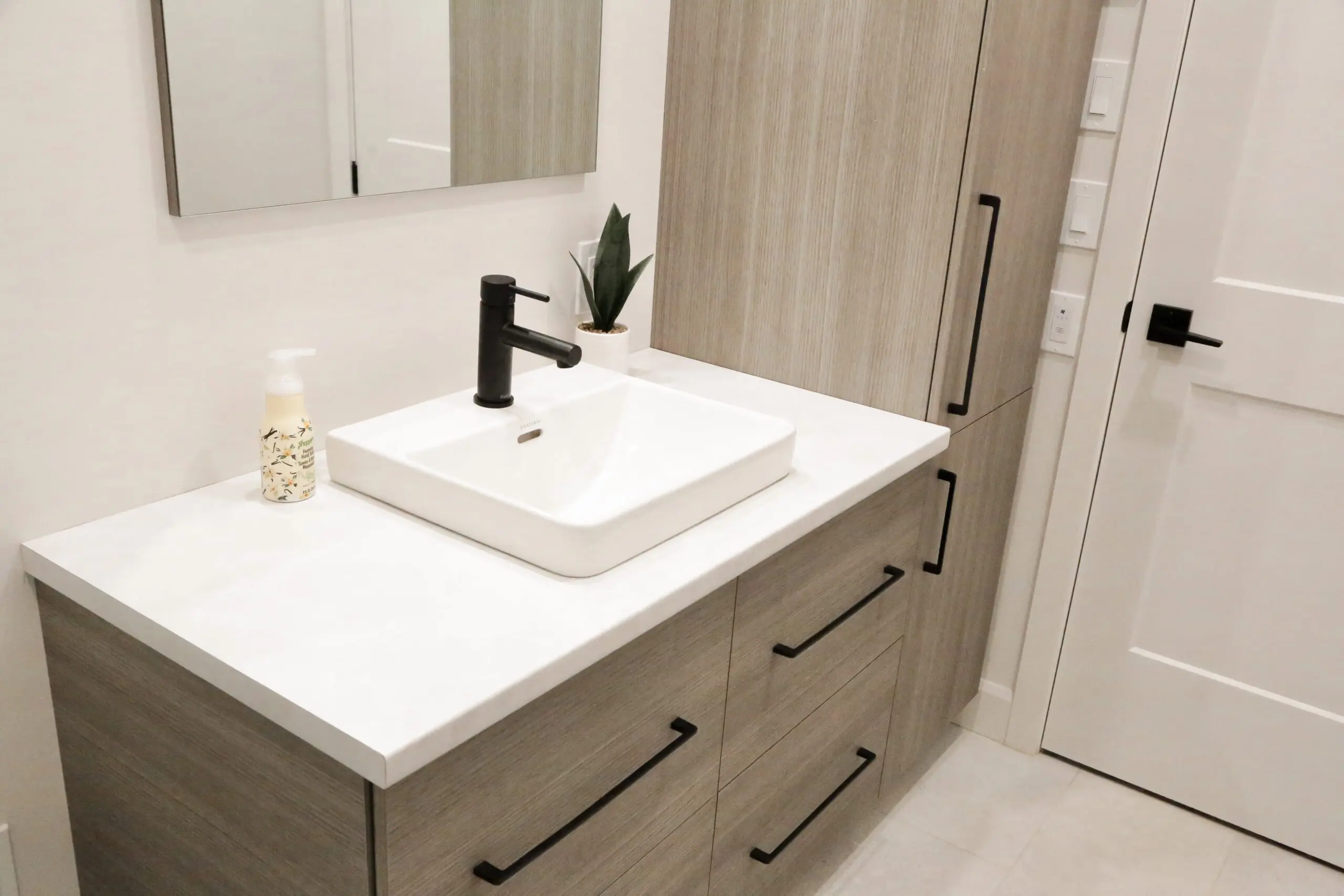 Meuble de salle de bain moderne avec un lavabo rectangulaire blanc, un robinet noir, un distributeur de savon, une petite plante, un grand miroir et des armoires en bois clair avec des poignées noires. La porte blanche et les murs clairs sont visibles.