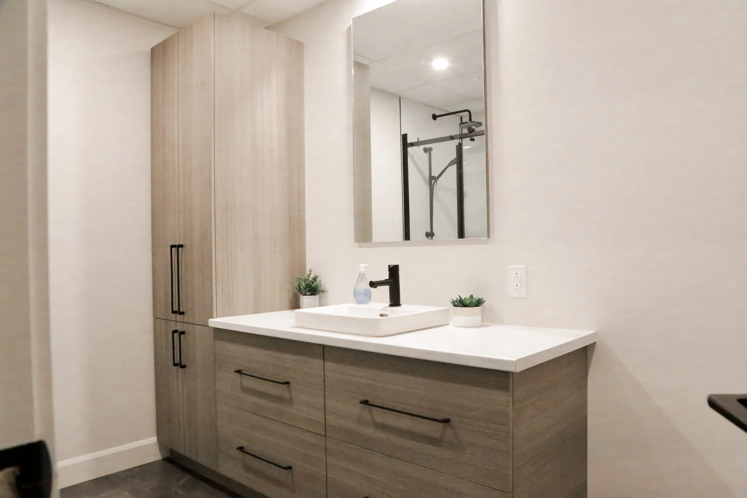 Salle de bains moderne avec des armoires en bois clair, un comptoir blanc, un lavabo rectangulaire avec un robinet noir, un miroir au-dessus du lavabo, un distributeur de savon, deux petites plantes en pot et un grand meuble de rangement sur la gauche. Douche avec porte en verre à l'arrière-plan.