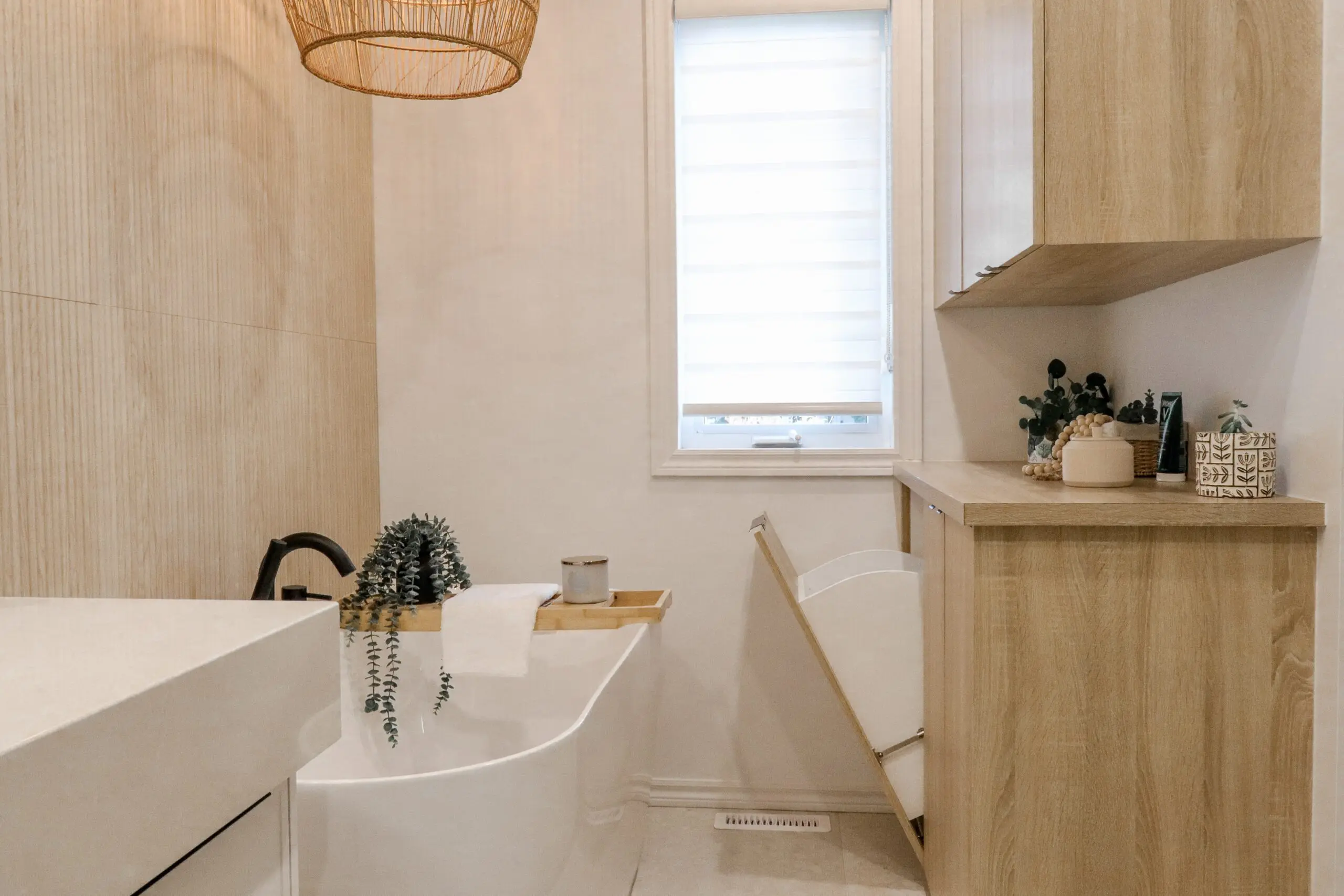 Salle de bains moderne avec une baignoire blanche autoportante, un plateau en bois, une serviette et une plante verte. Des armoires en bois clair, un panier à linge et une fenêtre avec un abat-jour blanc ajoutent au design épuré et minimaliste.