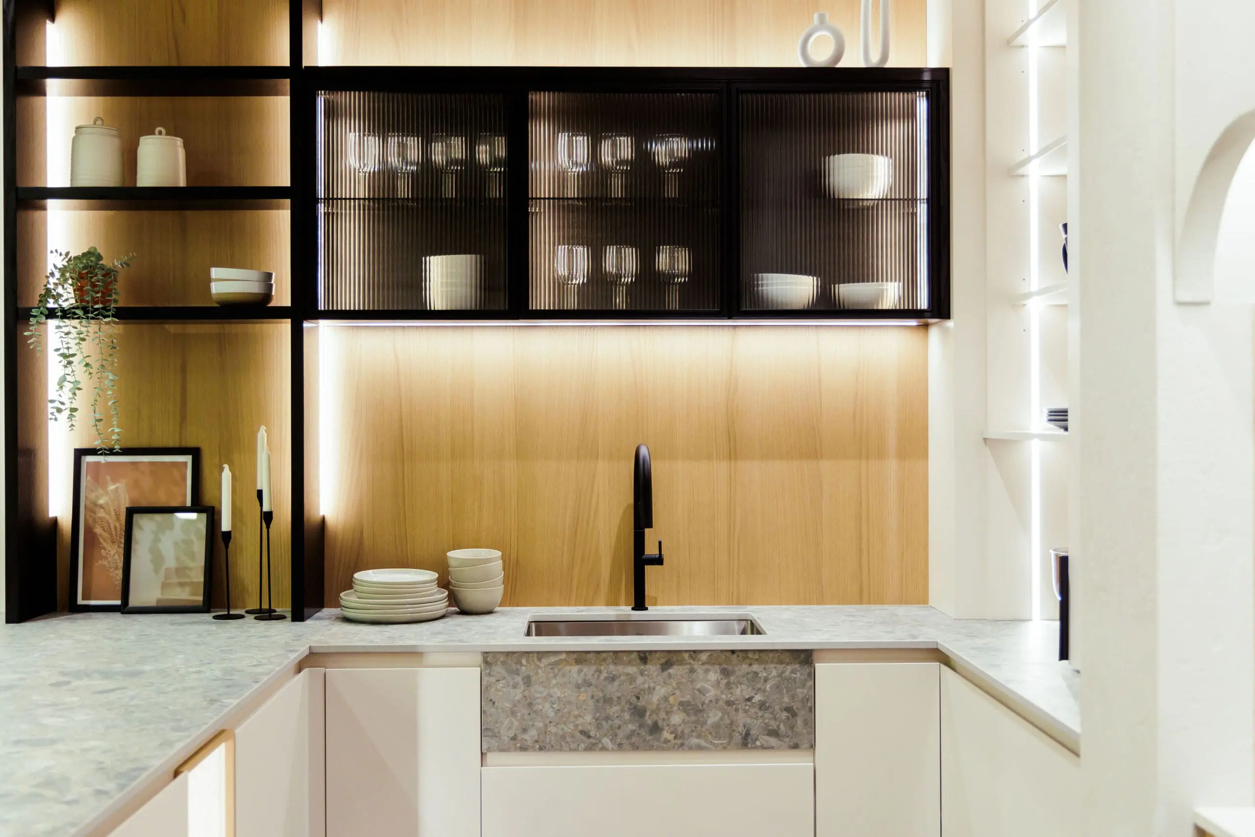 Cuisine moderne avec des meubles en bois clair et blanc, un plan de travail en pierre grise, un robinet noir et des étagères ouvertes. Les verres et les bols sont soigneusement rangés derrière les portes vitrées des armoires, et les assiettes et les bols sont empilés à côté de l'évier.