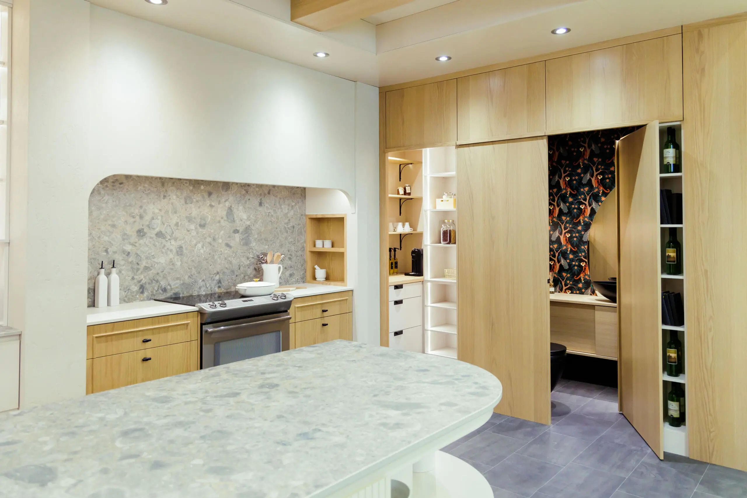 Cuisine moderne avec armoires en bois clair, comptoirs en pierre grise, cuisinière encastrée, étagères ouvertes et un garde-manger caché avec du papier peint floral, un éclairage élégant et un rangement organisé.