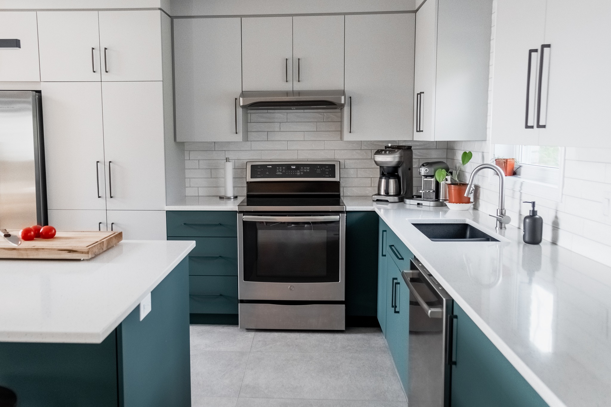 Cuisine moderne avec des armoires blanches et sarcelles, des appareils électroménagers en acier inoxydable, un évier noir et un comptoir avec une planche à découper, des tomates et des gadgets de cuisine. Dosseret en carreaux de faïence et lumière naturelle provenant d'une fenêtre.