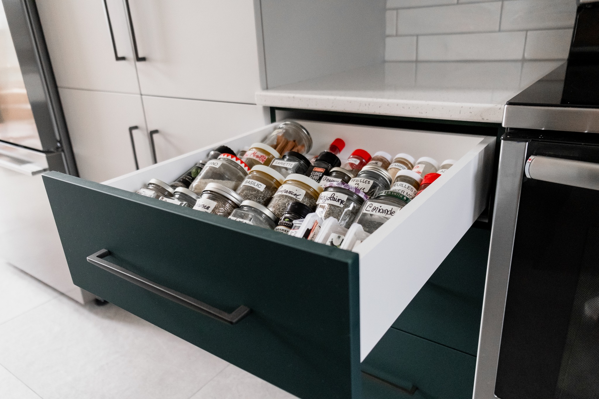 Un tiroir de cuisine ouvert, soigneusement organisé avec divers pots d'épices, chacun étiqueté et disposé en rangées. Le tiroir fait partie d'une cuisine moderne avec des armoires vert foncé et un comptoir blanc.