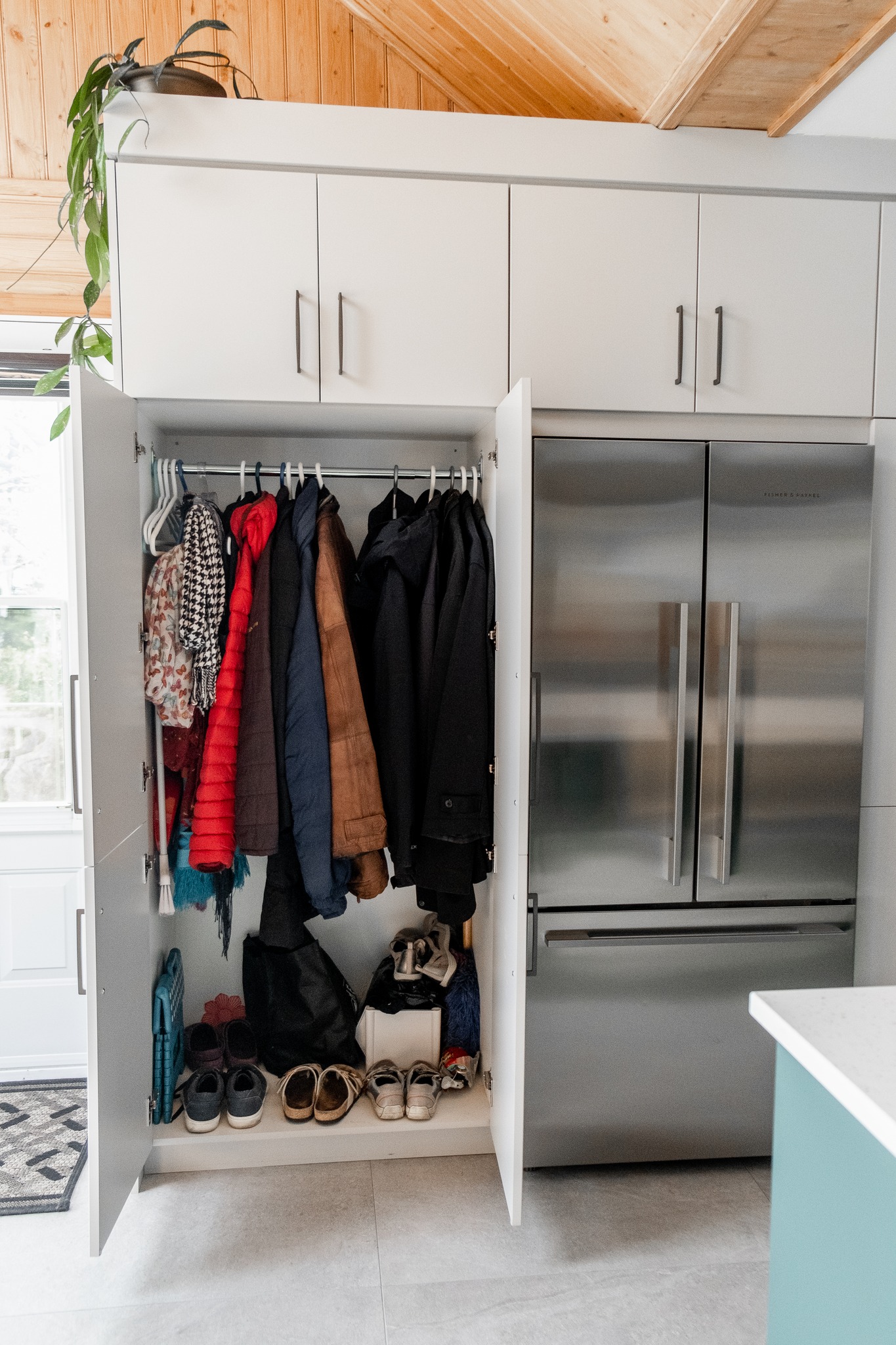 Placard ouvert à côté d'un réfrigérateur en acier inoxydable, montrant des manteaux sur des cintres, des écharpes, des chapeaux, des sacs sur l'étagère et plusieurs paires de chaussures bien rangées au fond. Une plante est posée sur le dessus de l'armoire.