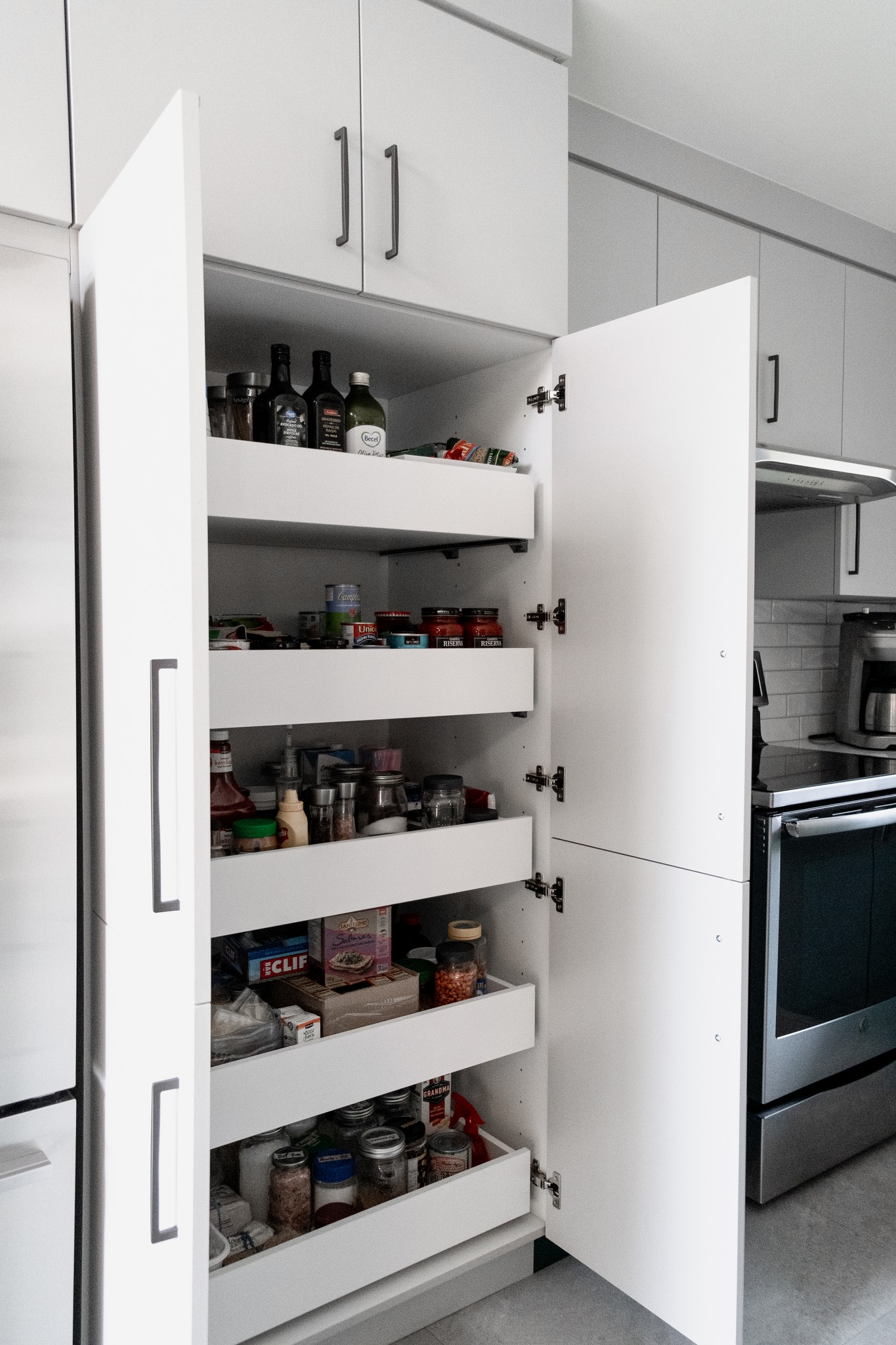 Un garde-manger de cuisine moderne avec des armoires blanches, ouvertes pour révéler des étagères organisées, remplies de bocaux, de boîtes de conserve, de sauces et de divers produits alimentaires. À proximité se trouvent des appareils électroménagers en acier inoxydable et d'autres armoires.