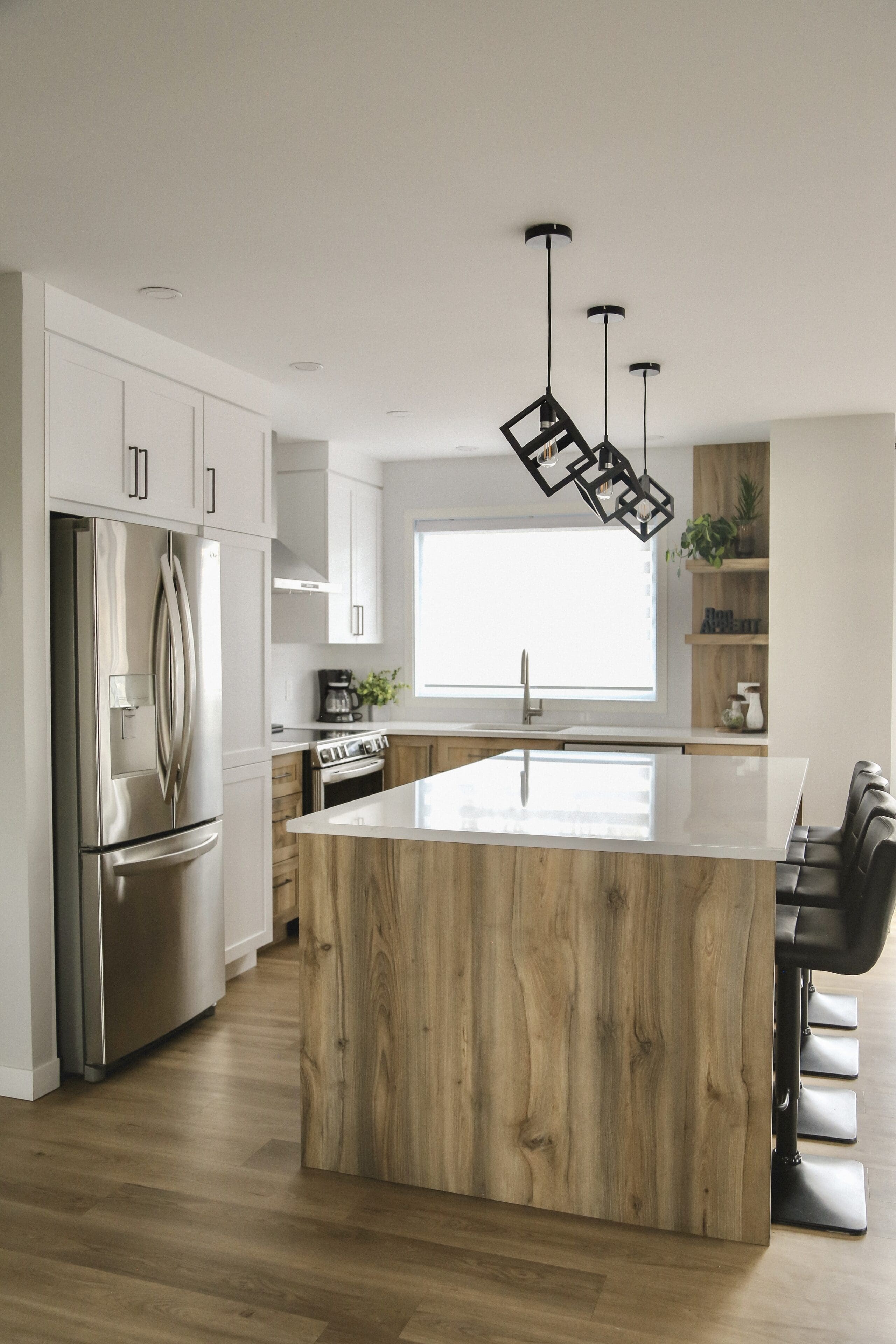 Cuisine moderne avec un grand îlot en bois, des armoires blanches, des appareils en acier inoxydable, des lampes suspendues noires, des tabourets de bar et une grande fenêtre laissant entrer la lumière naturelle.