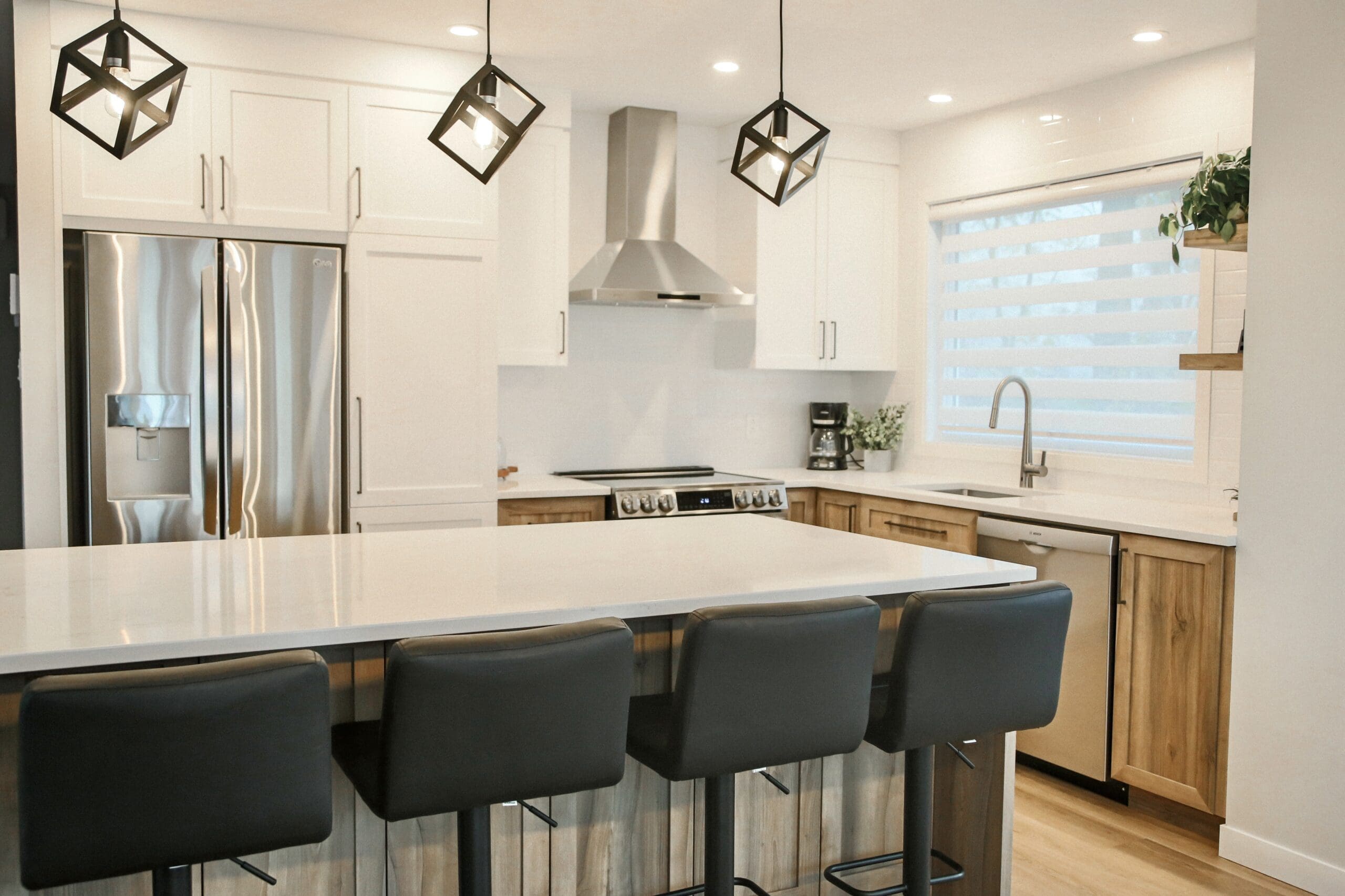 Cuisine moderne avec des armoires blanches, des appareils en acier inoxydable, un îlot central avec quatre tabourets de bar noirs, des lampes suspendues géométriques, une grande fenêtre avec des stores et des accents en bois sur les armoires inférieures.