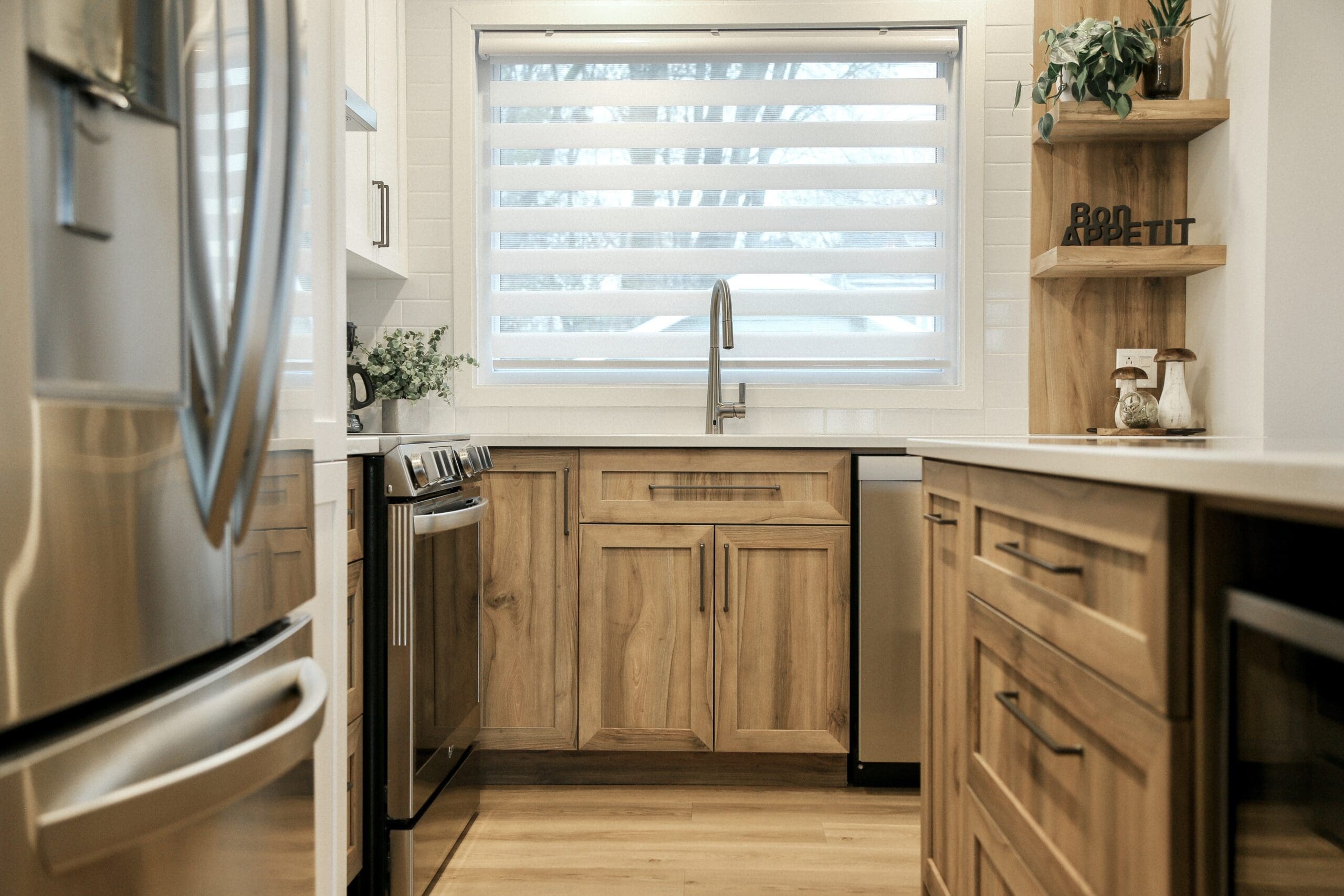 Cuisine moderne avec des armoires en bois clair, des appareils en acier inoxydable, une grande fenêtre avec des stores, un robinet à col de cygne, des comptoirs blancs et des plantes comme décor sur le comptoir et l'étagère.