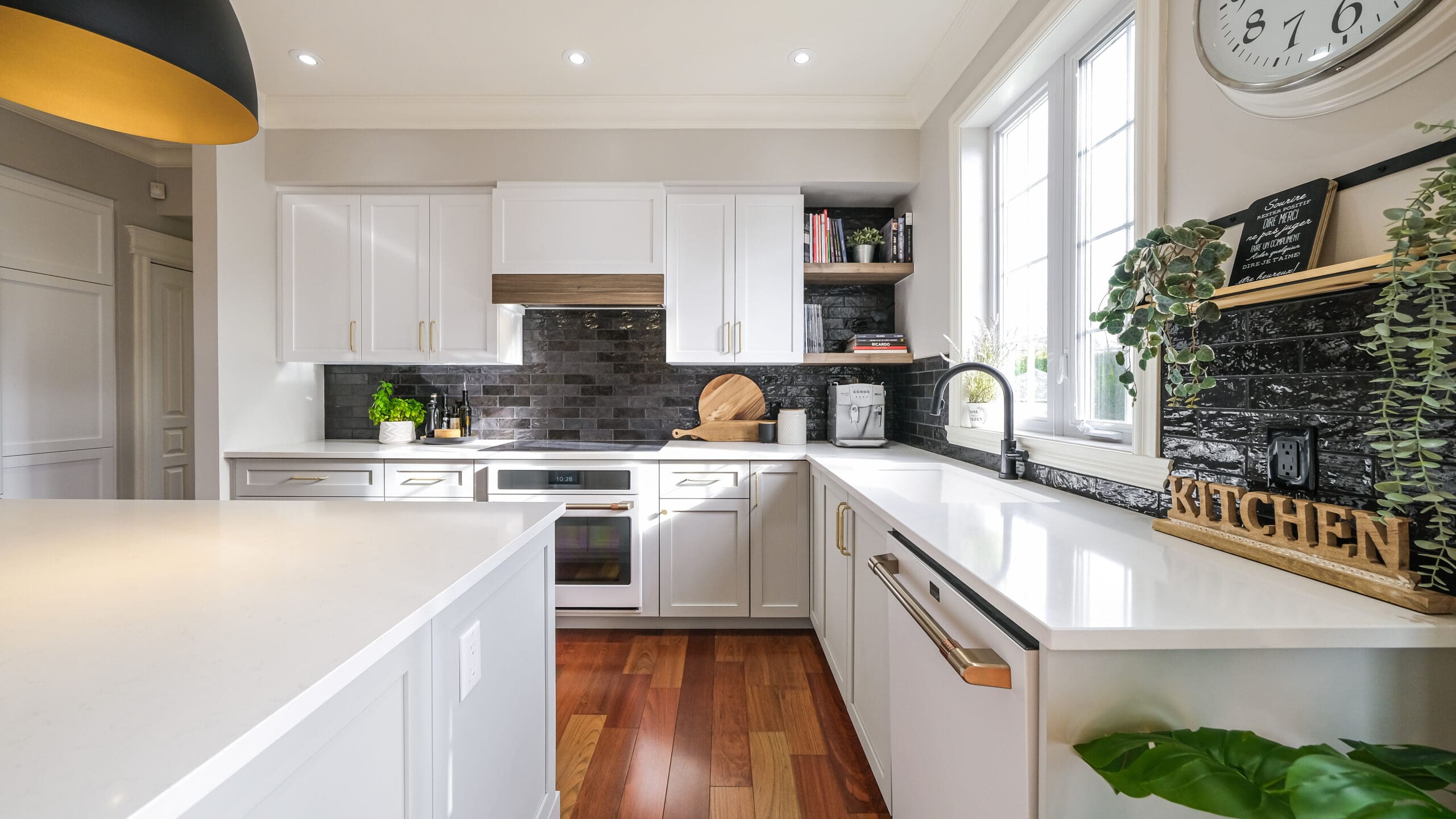 Cuisine moderne avec des armoires blanches, un dosseret en carreaux noirs, des appareils en acier inoxydable, un plancher en bois, de grandes fenêtres, des plantes décoratives et des ustensiles de cuisine sur les comptoirs et les étagères. Espace lumineux, propre et organisé.