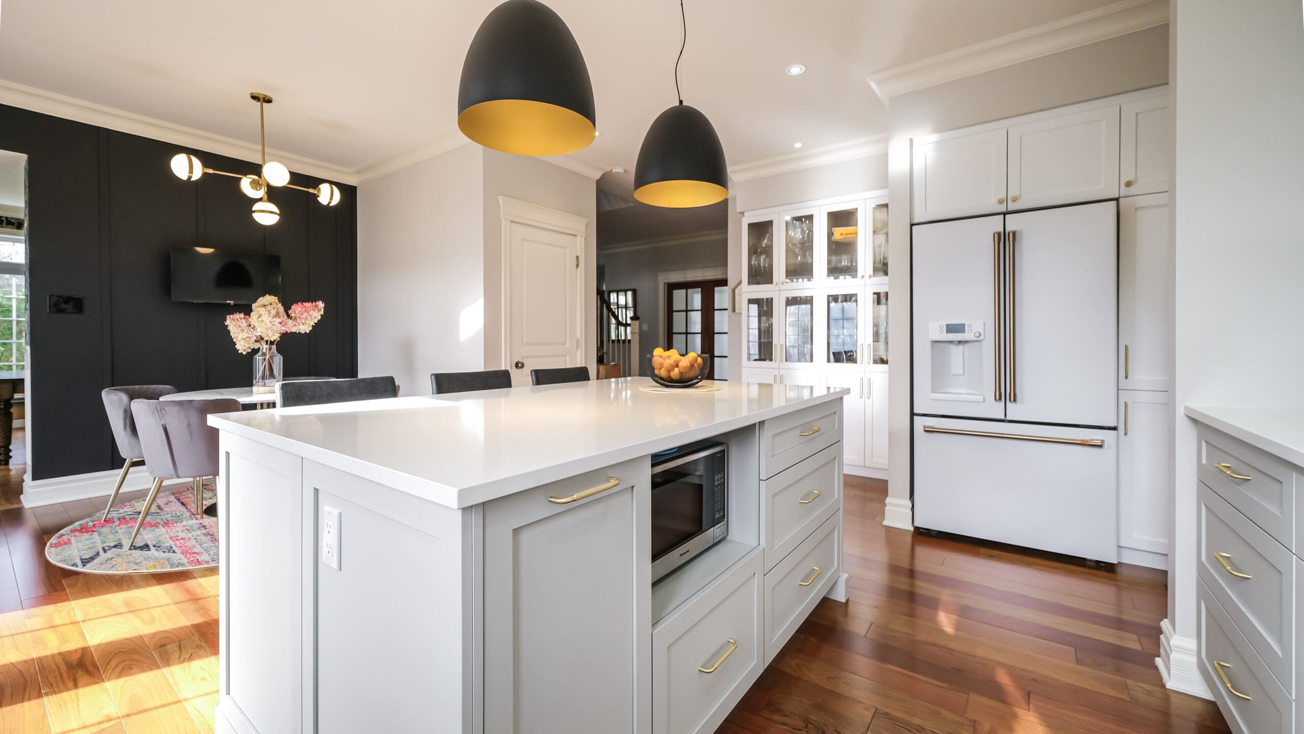 Cuisine moderne avec un grand îlot blanc, des luminaires suspendus aux accents dorés, des armoires blanches, un four à micro-ondes encastré, du parquet et un coin repas visible à l'arrière-plan. Un bol de citrons est posé sur l'îlot.