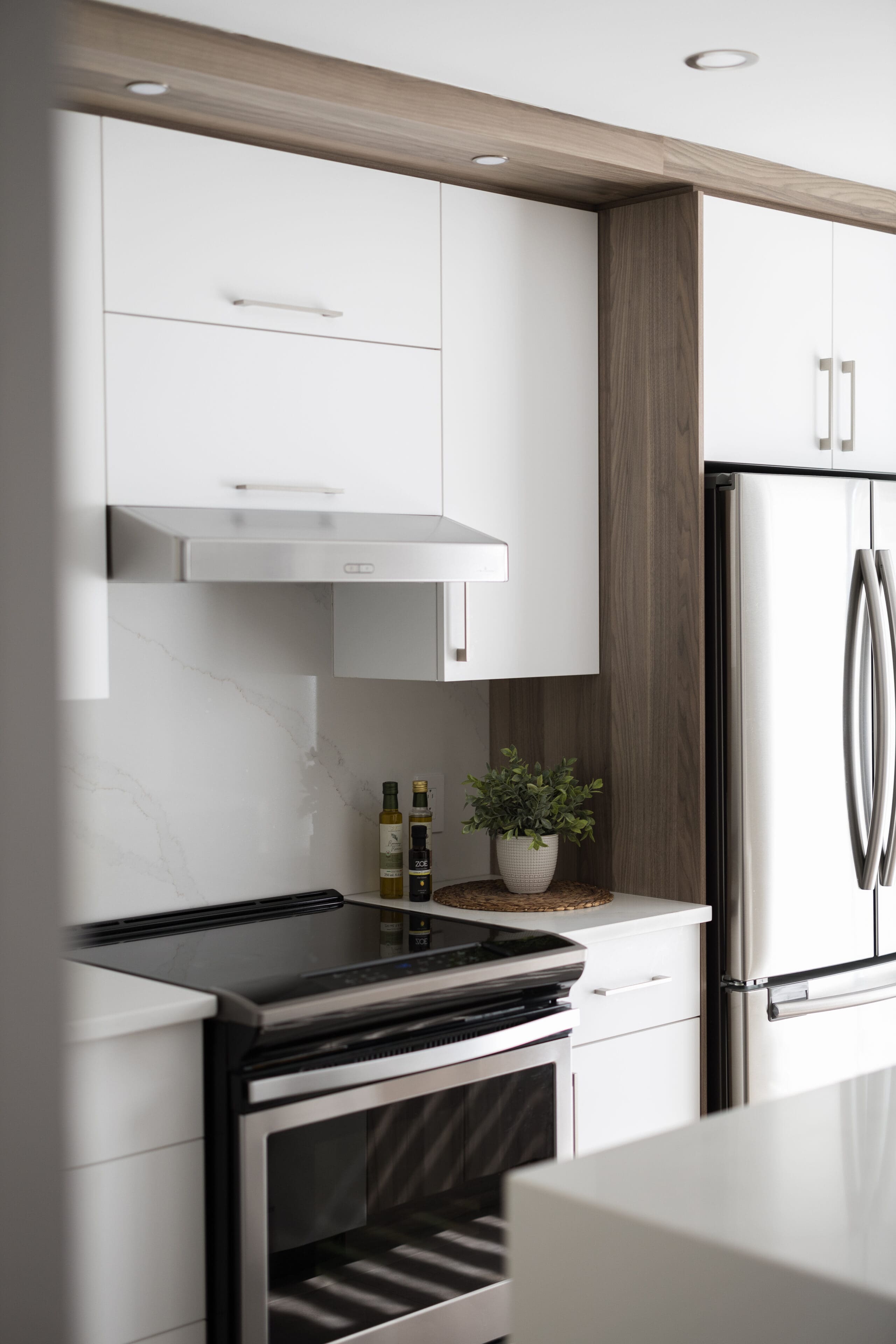 Cuisine moderne avec des armoires blanches, un four et une hotte en acier inoxydable, un réfrigérateur argenté à double porte, une plante en pot et des bouteilles d'huile sur un comptoir blanc avec un dosseret en marbre.