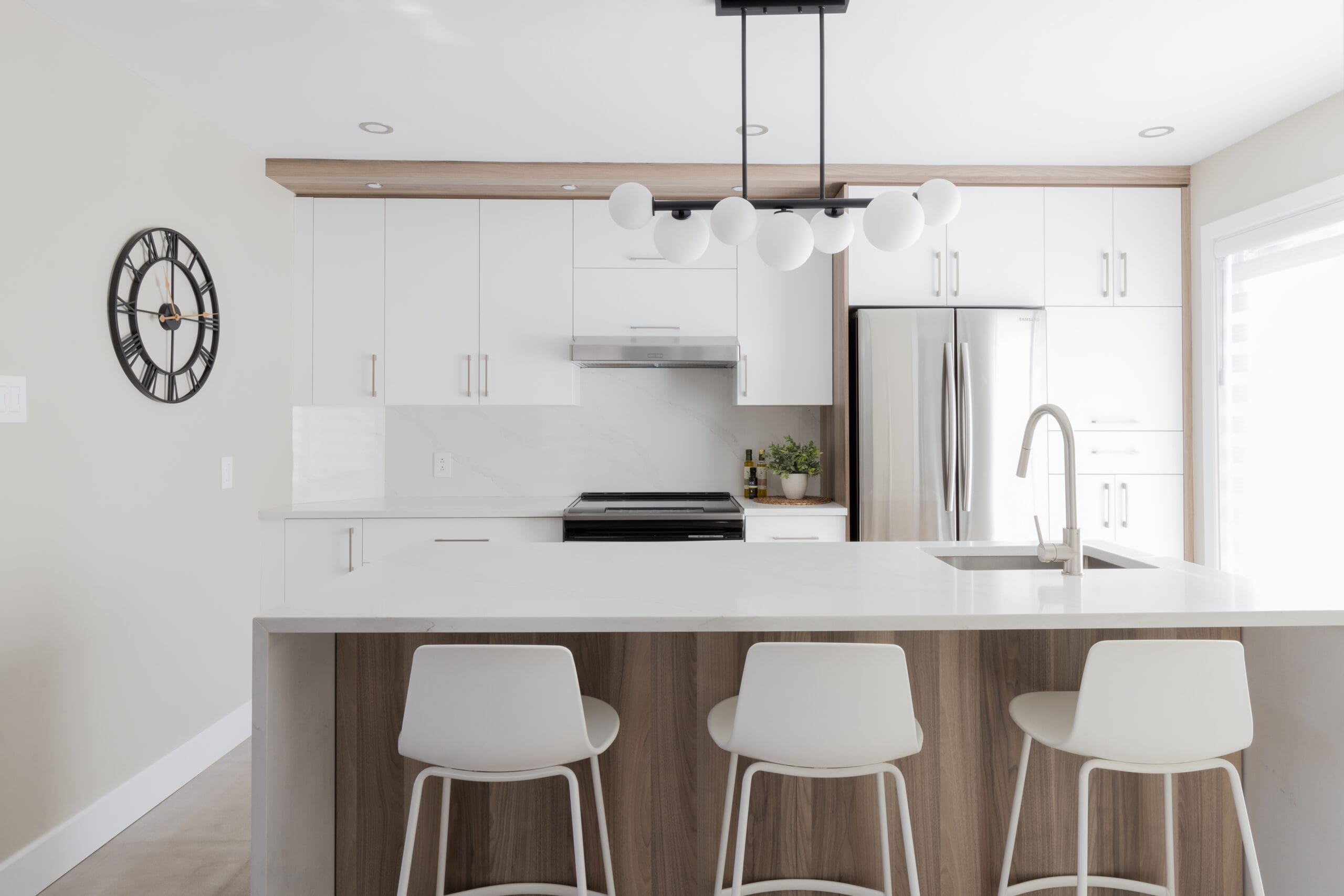 Cuisine moderne avec des armoires blanches, un réfrigérateur en acier inoxydable, un grand îlot avec trois chaises blanches, une horloge minimaliste en noir et blanc sur le mur et un luminaire contemporain au-dessus de l'îlot.