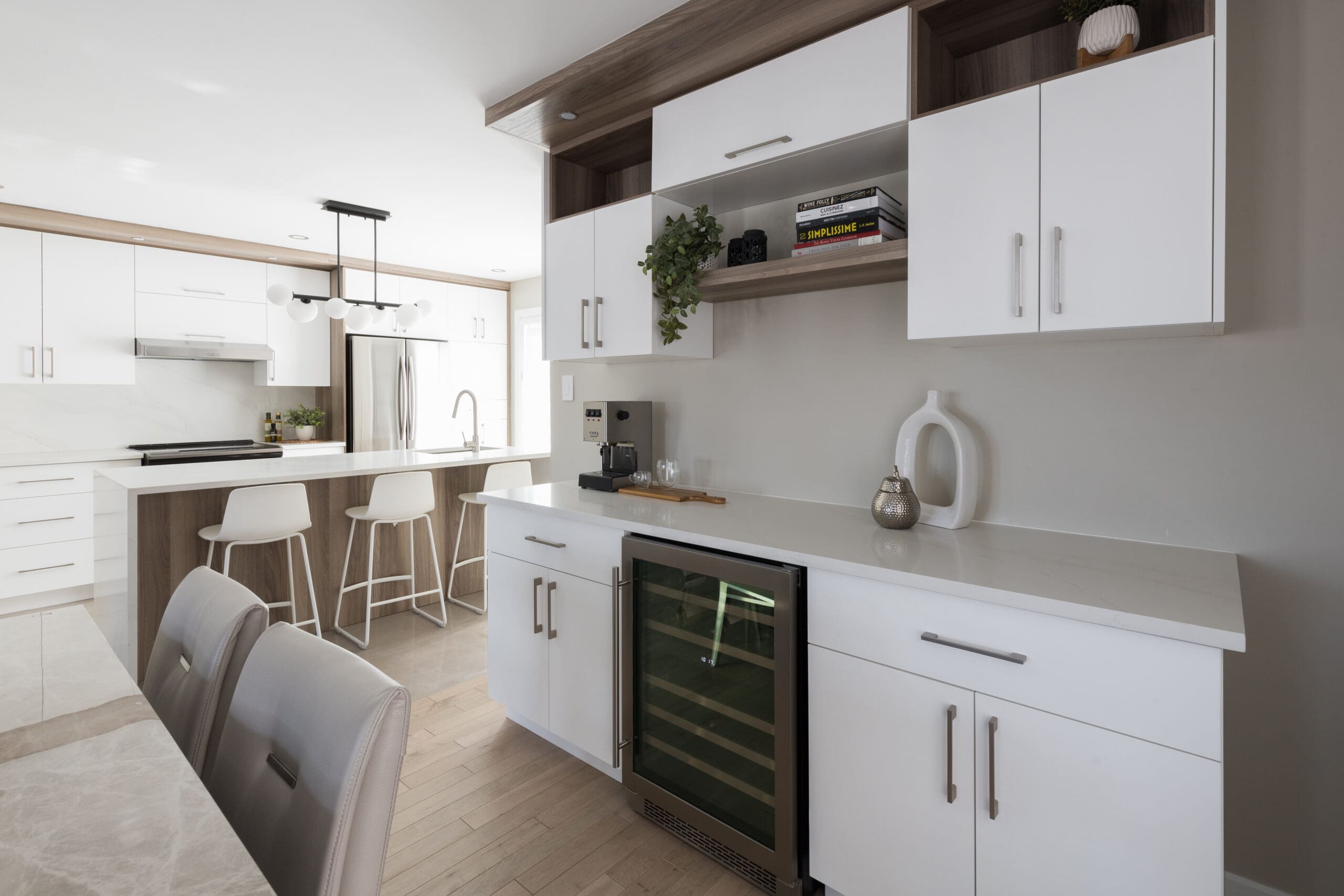 Cuisine moderne avec des armoires blanches, un réfrigérateur à vin, des étagères ouvertes, une cafetière et des tabourets de bar sur un îlot. Des plantes décoratives et des livres ajoutent des accents à cet espace clair et épuré.
