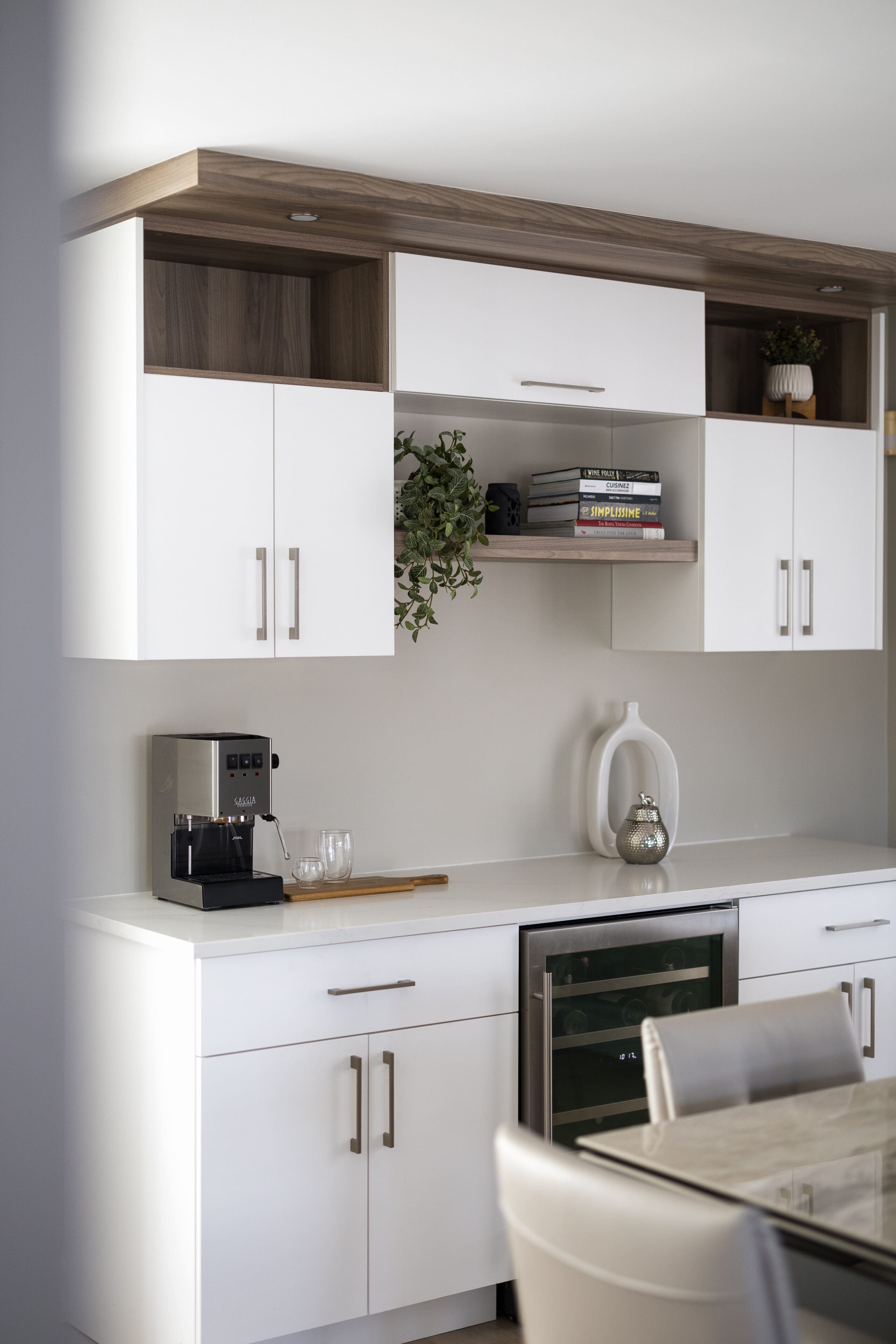 Cuisine moderne avec des armoires blanches, un comptoir sur lequel se trouvent une machine à café, deux verres, un plateau en bois, un vase décoratif et quelques plantes, avec des livres et des objets de décoration sur des étagères ouvertes au-dessus.
