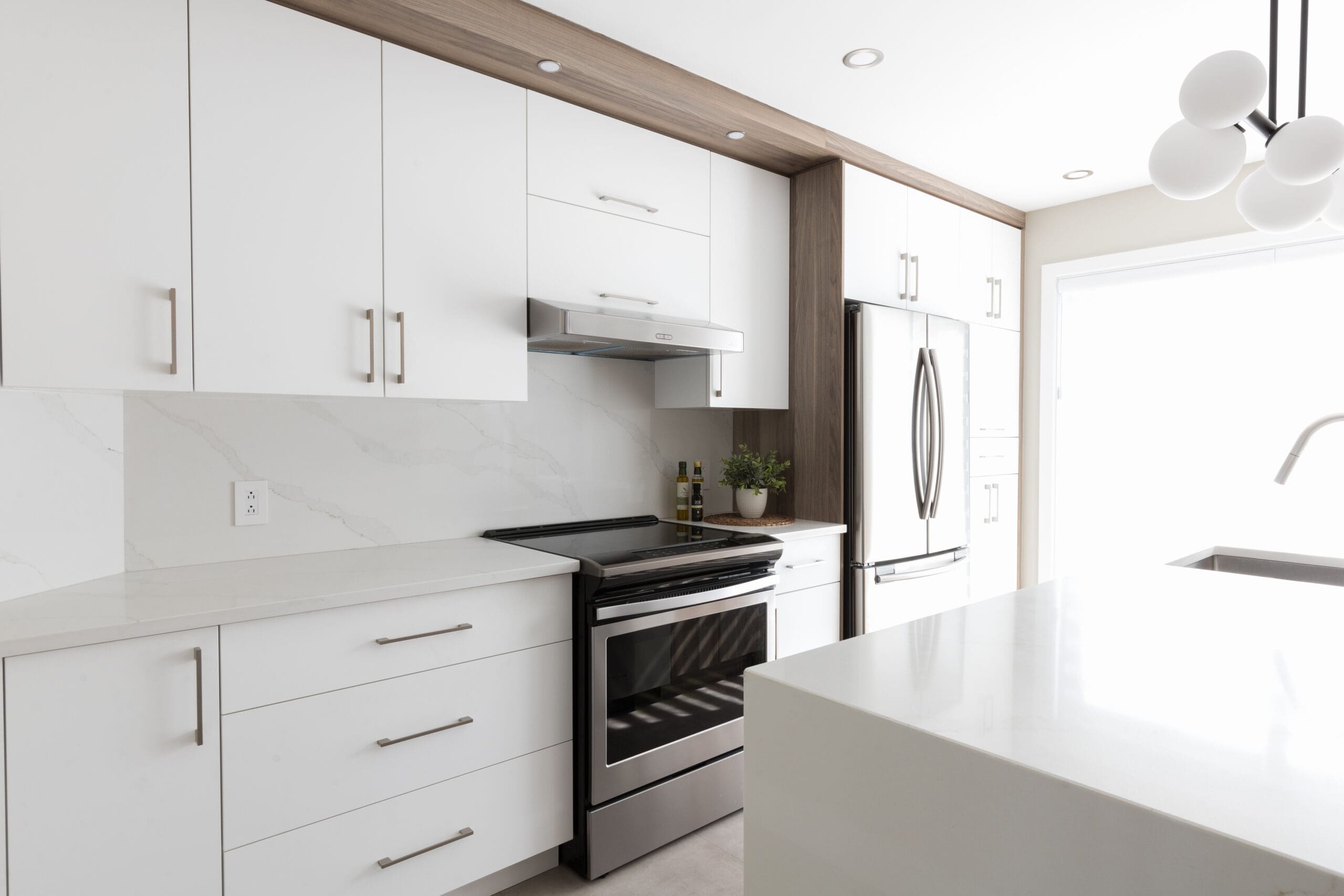 Cuisine moderne avec des armoires blanches, des appareils en acier inoxydable, un dosseret en marbre et un îlot. Une cuisinière avec une hotte est au centre, et un réfrigérateur se trouve à droite, près d'une grande fenêtre.