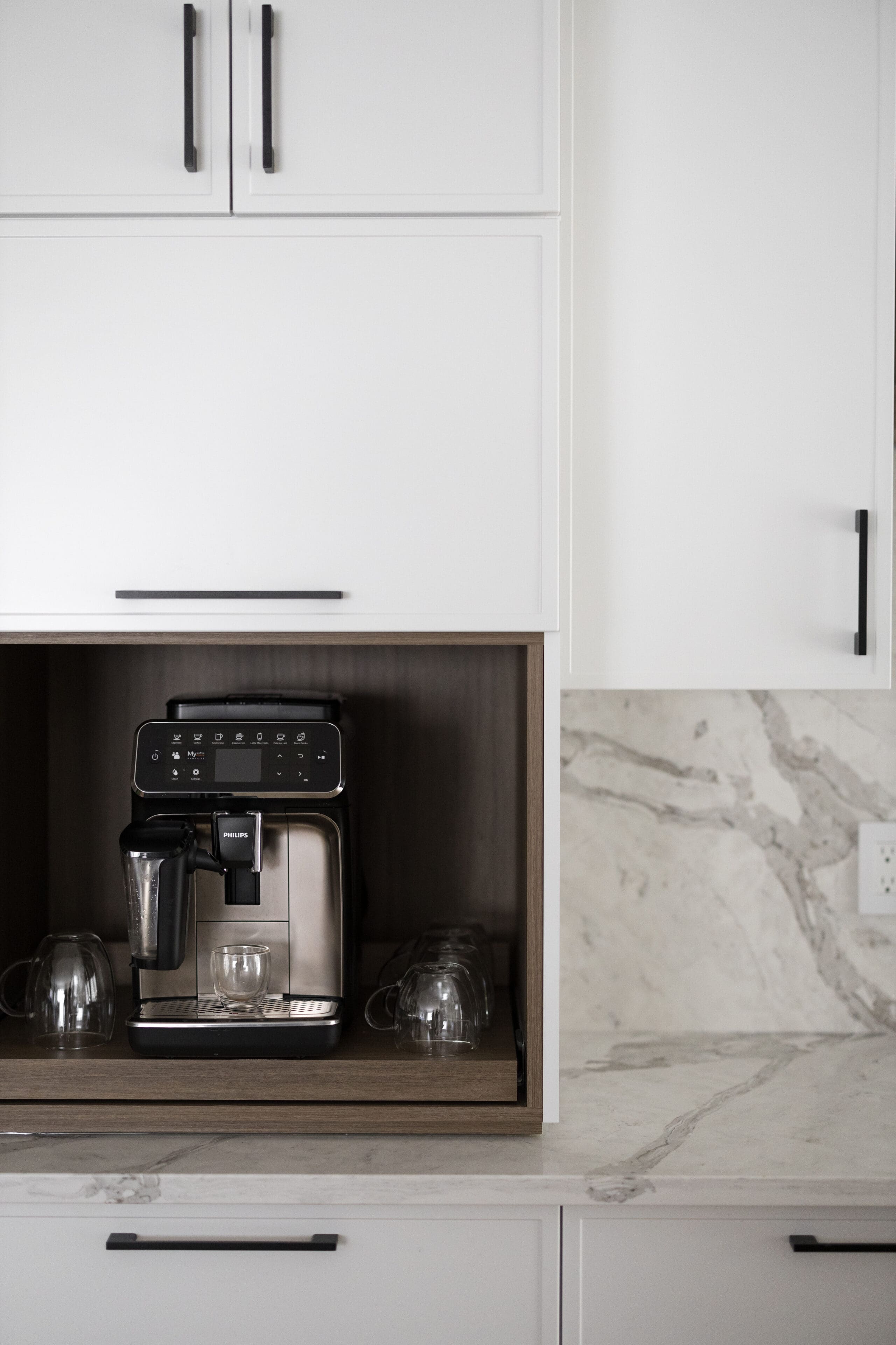 Une machine à café moderne avec des tasses en verre est placée dans un coin encastré dans une cuisine blanche avec des comptoirs en marbre et des poignées d'armoire foncées.