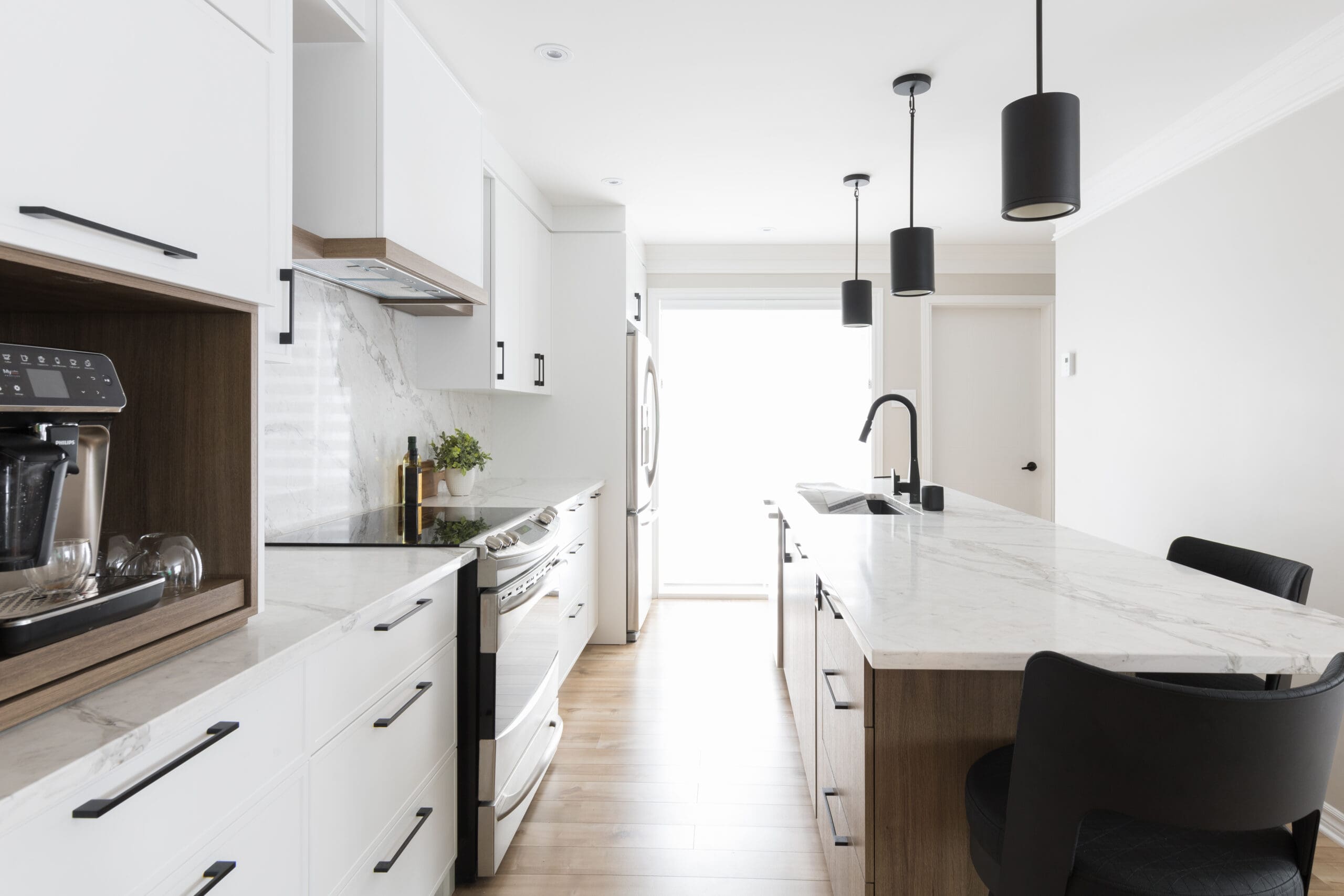 Cuisine moderne avec armoires blanches, comptoirs en marbre, appareils électroménagers en acier inoxydable, cafetière intégrée, lampes suspendues noires, grand îlot avec évier et chaises noires sur un sol en bois clair.