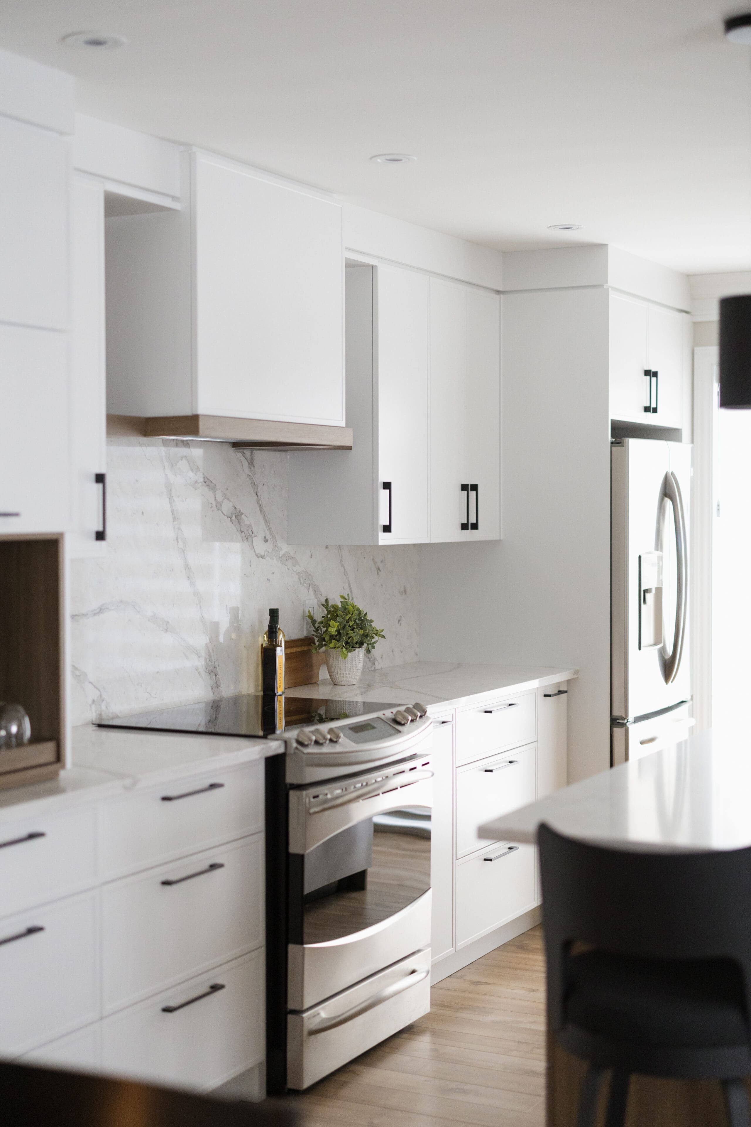 Cuisine moderne avec des armoires blanches, des appareils en acier inoxydable, un dosseret en marbre, une plante en pot, une bouteille d'huile sur le comptoir et un sol en bois. L'espace est baigné de lumière naturelle.
